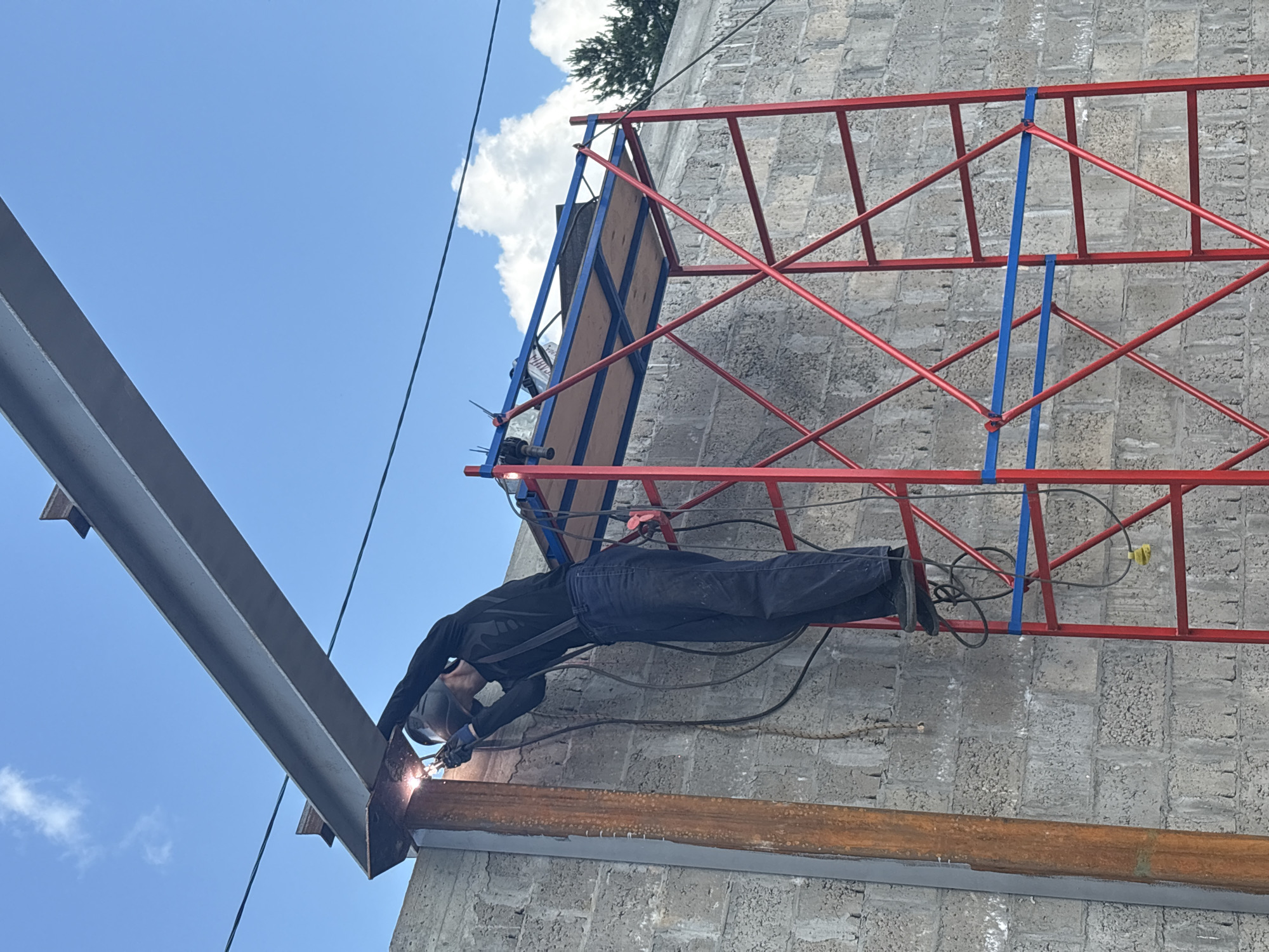 A male construction worker, wearing a black long-sleeved shirt, dark pants, suspenders, safety glasses, and gloves, is welding a large steel beam to a vertical steel support. He is standing on a red and blue metal scaffold. The background is a gray concrete block wall and a bright blue sky with some white clouds. A tall, rusty metal pole is visible to the left of the worker. The scene appears to be an outdoor construction site, possibly in the city of Chaikovsky, Russia, given the presence of a Russian flag on the scaffolding.