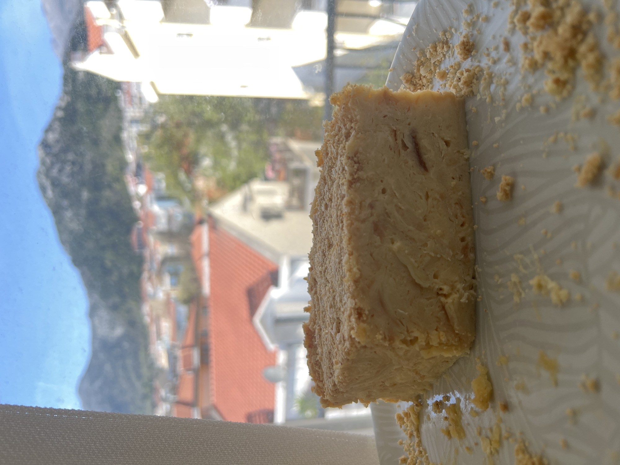 A slice of cake rests on a white plate with scattered crumbs, likely a graham cracker crust, in front of a window. The background reveals a partial cityscape with buildings featuring red-tiled roofs, green vegetation, and a distant mountainous area meeting a blue sky. This scene is likely in Budva, Montenegro, given the architectural style and geographic features. The image captures a still life composition of food with a scenic urban backdrop.