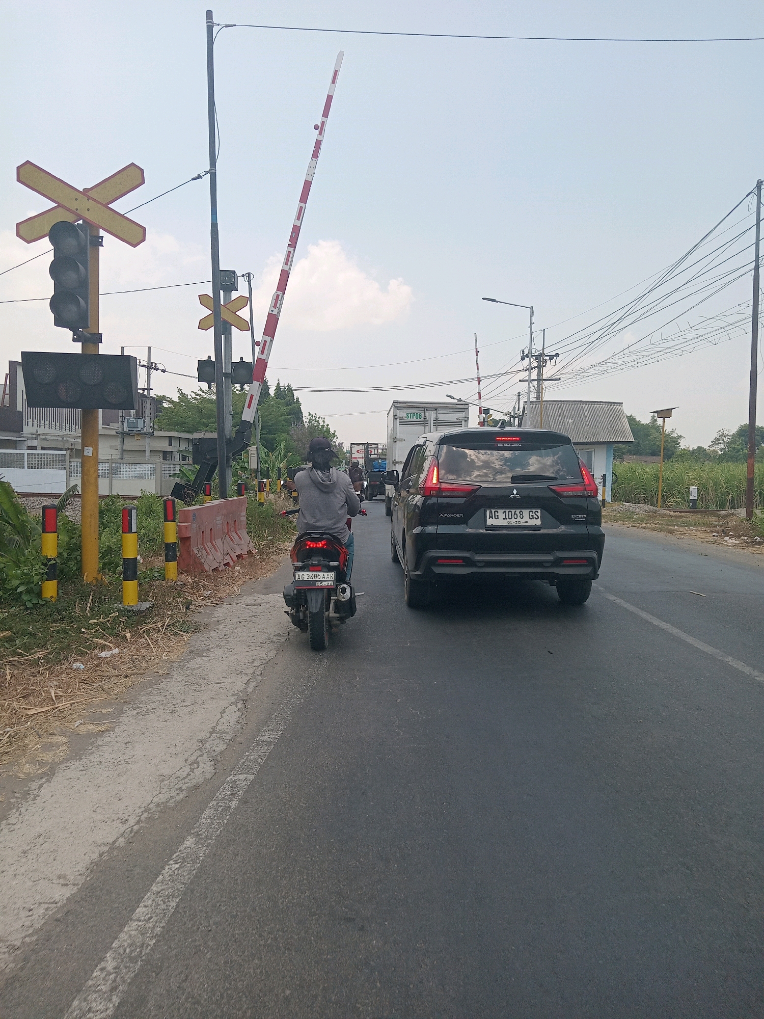 This is a photograph of a railroad crossing in Branggahan, Indonesia. The crossing gate is partially lowered, indicating that a train may be approaching or passing. A black Mitsubishi Xpander SUV with license plate AG 1068 GS is stopped on the road, waiting for the gate to open. A person on a red scooter is also stopped behind the SUV, facing away from the camera. The road is asphalt, with a white line marking the lane. To the left of the road is a railroad signal with red, yellow, and black lights, and a yellow railroad crossing sign. There are also several striped yellow and black bollards along the side of the road. In the background, there are houses, trees, and power lines. The sky is overcast.