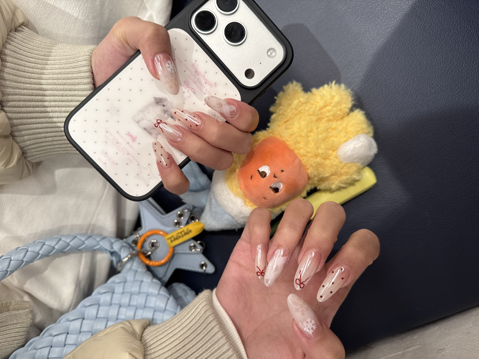 The image presents a close-up, top-down perspective of an individual's hands and personal accessories, situated indoors in Yuzhong District, China. The person is wearing a light-colored, ribbed long-sleeved garment and a beige puffed jacket. Both hands display long, almond-shaped fingernails, each featuring a clear or translucent base with distinct decorative patterns. These designs include red polka dots, small red bows, and white snowflake motifs.
The right hand holds a white-cased iPhone, identifiable by its triple-lens camera system on the rear, with the case displaying faint pink dot patterns. Adjacent to the left hand, resting on a dark blue, smooth surface, are several items. These include a light blue braided handbag with a metallic clasp, to which a blue star-shaped charm with silver studs is attached. Also connected to the handbag is an orange key-shaped keychain bearing black Chinese characters that translate to "Yuzhong District." Next to the individual's left hand lies a yellow, fuzzy plush toy with an orange head, small black eyes, and white stubby feet.