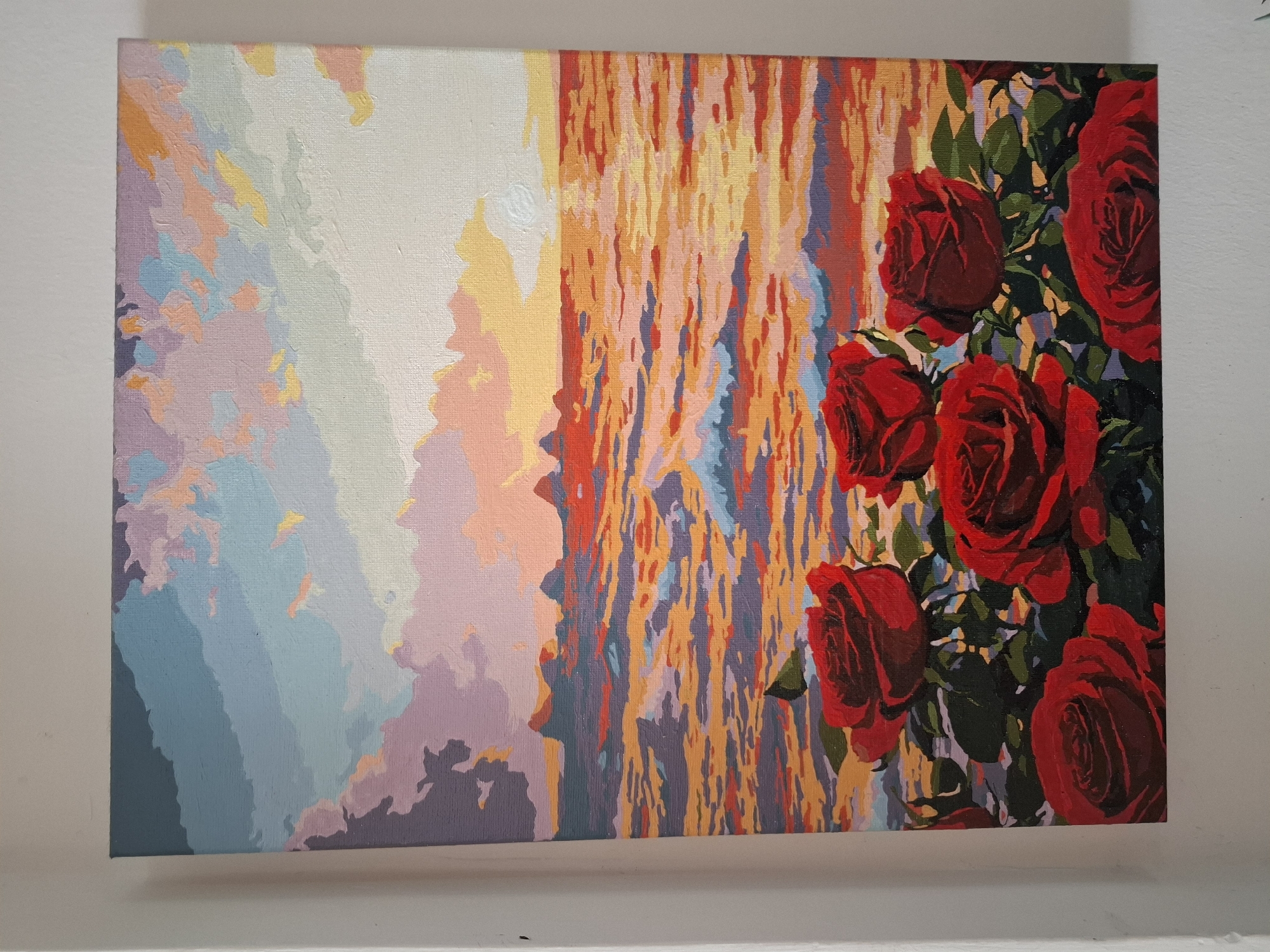 The image displays a painting with a seascape at sunset, foregrounded by red roses. The sky is rendered in soft hues of blue, pink, purple, and yellow, with streaks of orange and white indicating the setting sun. The sea reflects these colors, appearing as a vibrant tapestry of reds, oranges, and blues. Jagged, dark shapes, possibly islands or rock formations, punctuate the horizon. The roses in the foreground are a deep, rich red, with dark green leaves and stems. The painting's style is impressionistic, with visible brushstrokes creating a textured effect. There is no indication of any specific location in Nha Trang, Vietnam, within the image.