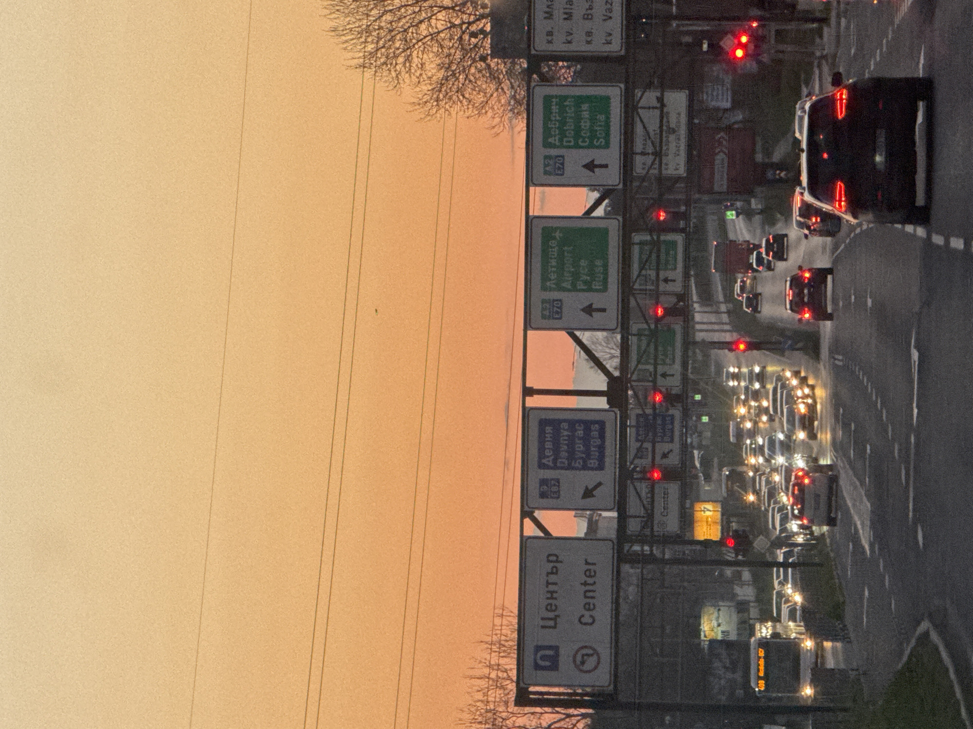 The image shows a section of a highway in what appears to be Varna, Bulgaria. The view is along a road with multiple lanes occupied by dense traffic, with many cars stopped at a red light. The photograph seems to have been taken at dusk or dawn, as the sky is a soft orange. Along the roadside are several green directional signs indicating various destinations, including "Center," "Devnya," "Burgas," "Airport," "Ruse," "Dobrich," and "Sofia." The signs also indicate the European route numbers E70 and E87. There are street lights and overhead wires running parallel to the road. The photograph may have been taken from an elevated position, such as a building or bridge.