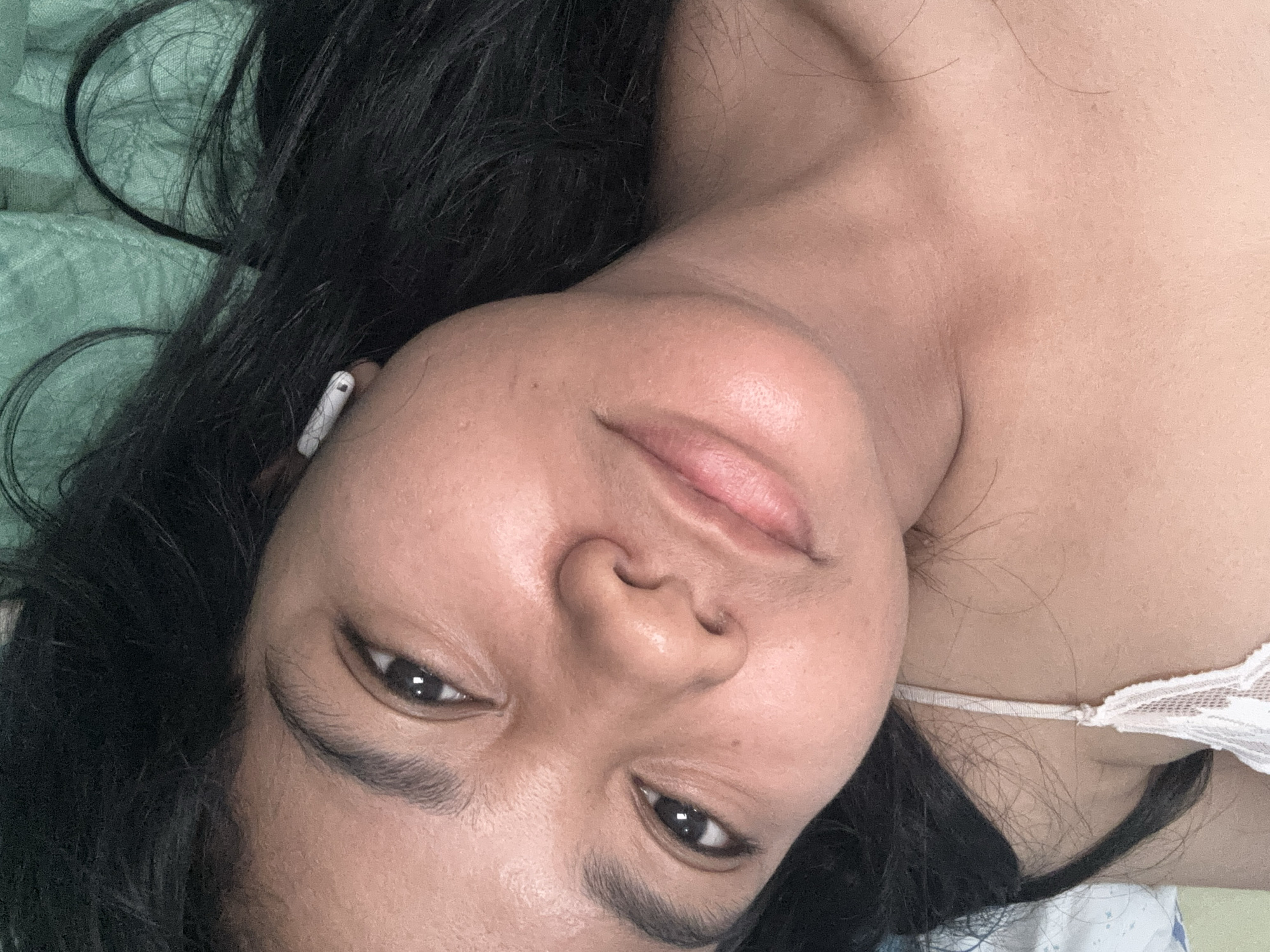 A close-up, overhead, selfie-style shot shows a young woman lying down, her head tilted to the side. Her dark hair is spread out around her face, and she has a white wireless earbud in her right ear. She is wearing a white camisole with lace trim. The background appears to be a bed with a teal green textured blanket. The image was taken in North Jakarta, Indonesia.