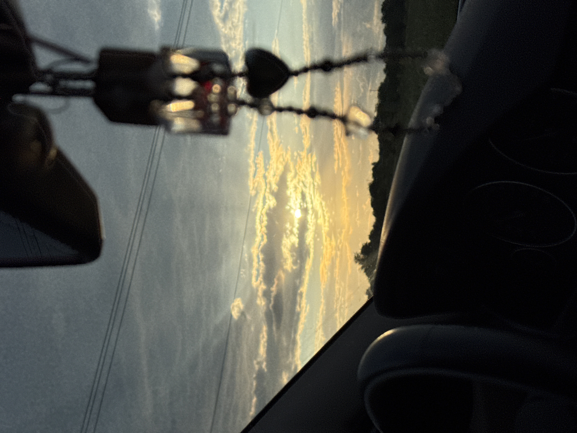 This media file depicts a view from inside a vehicle looking out the windshield during sunset. The sun is low in the sky, casting a warm, golden light on the clouds. The clouds are wispy and elongated, with streaks of light suggesting movement. The sky is a gradient of soft blues and oranges.
In the foreground, a beaded string with a heart-shaped charm is hanging from the rearview mirror. The beads are mostly dark, with some reflective elements. Part of the vehicle's interior is visible, including the dashboard and a headrest, rendered in dark shadows that contrast with the bright sky. A side mirror is also partially visible on the left.
Beyond the windshield, a landscape of fields and trees can be seen, silhouetted against the setting sun. In the distance, there appear to be power lines stretching across the horizon. The overall mood is serene and contemplative, likely captured during a drive. The time of day is clearly sunset, and the weather appears clear with scattered clouds. The location context of Novomar'evskaya, Russia, suggests a rural or semi-rural setting. No text is visible in the image.