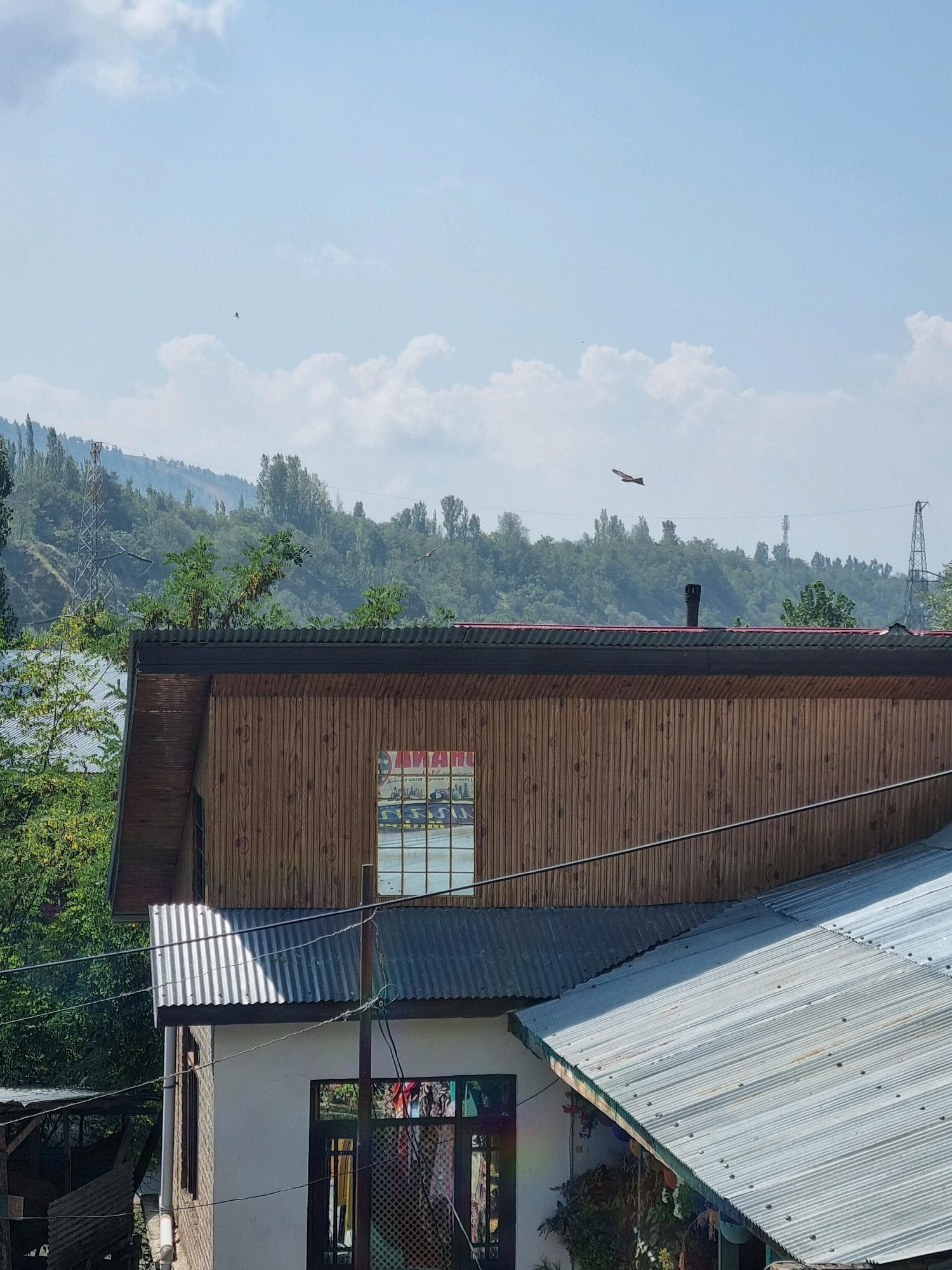 A medium shot captures a bright, sunny day in Manigam, India. The foreground features the tops of several buildings with corrugated metal roofs. One building, constructed with vertical wooden planks, has a visible window reflecting the sky and trees. A small black chimney protrudes from a red-tiled roof on this building. In the background, a lush, green hillside is covered in trees. A tall, silver electricity pylon stands on the left side of the hill, and another on the right. The sky is a clear blue with scattered white clouds. A bird is in flight in the upper portion of the frame. The overall scene depicts a rural or semi-rural setting.