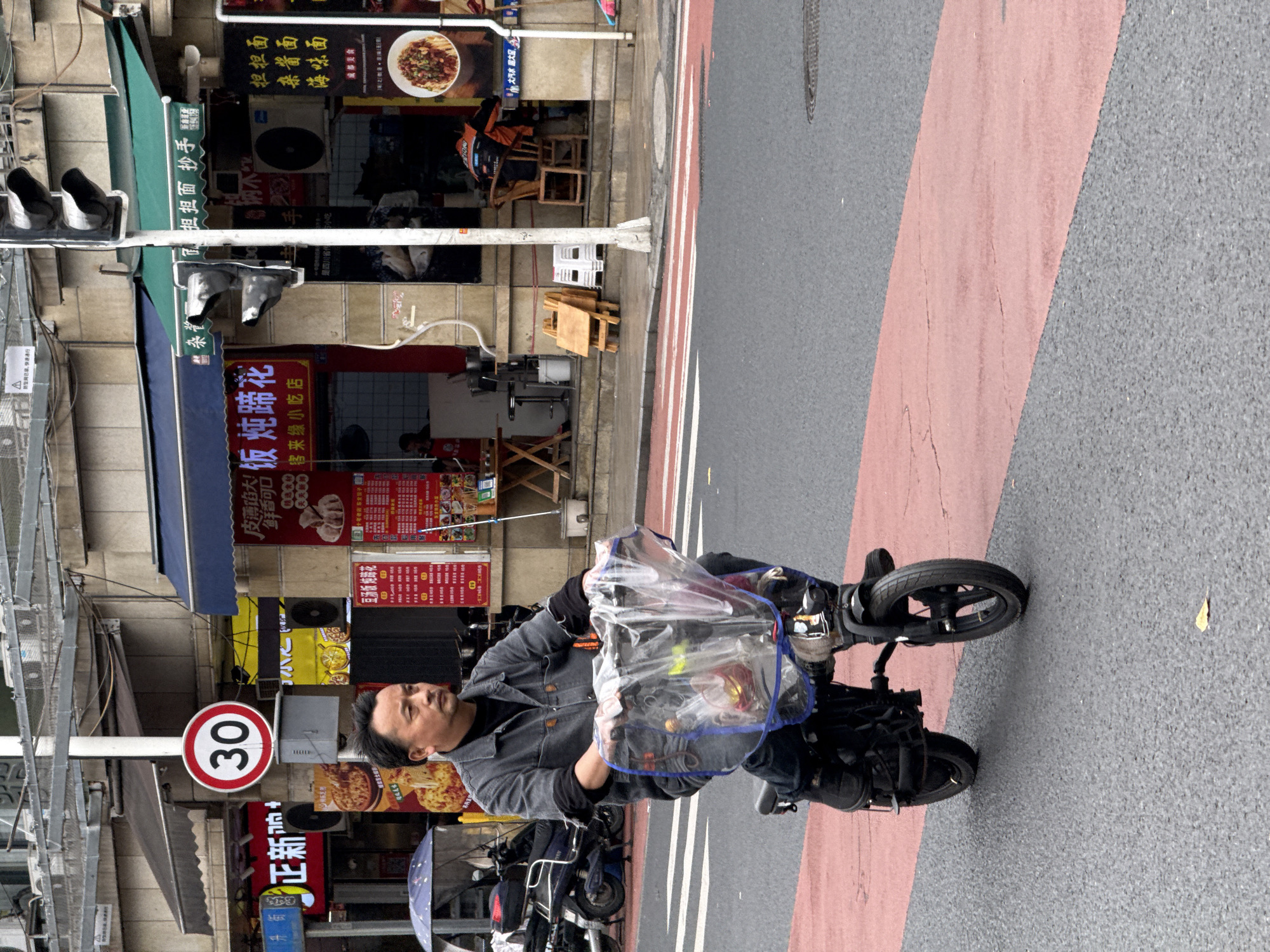 The image captures a man on a small electric scooter in the Jinjiang District of Chengdu, China. He is dressed in a denim jacket and black shirt, and carries a plastic bag filled with items on the front of his scooter. The scene is an urban environment with Chinese storefronts, signs, and traffic signals visible in the background. There is a speed limit sign indicating "30" nearby. The street has grey pavement and a reddish-pink painted bike lane. Other scooters are parked along the sidewalk.