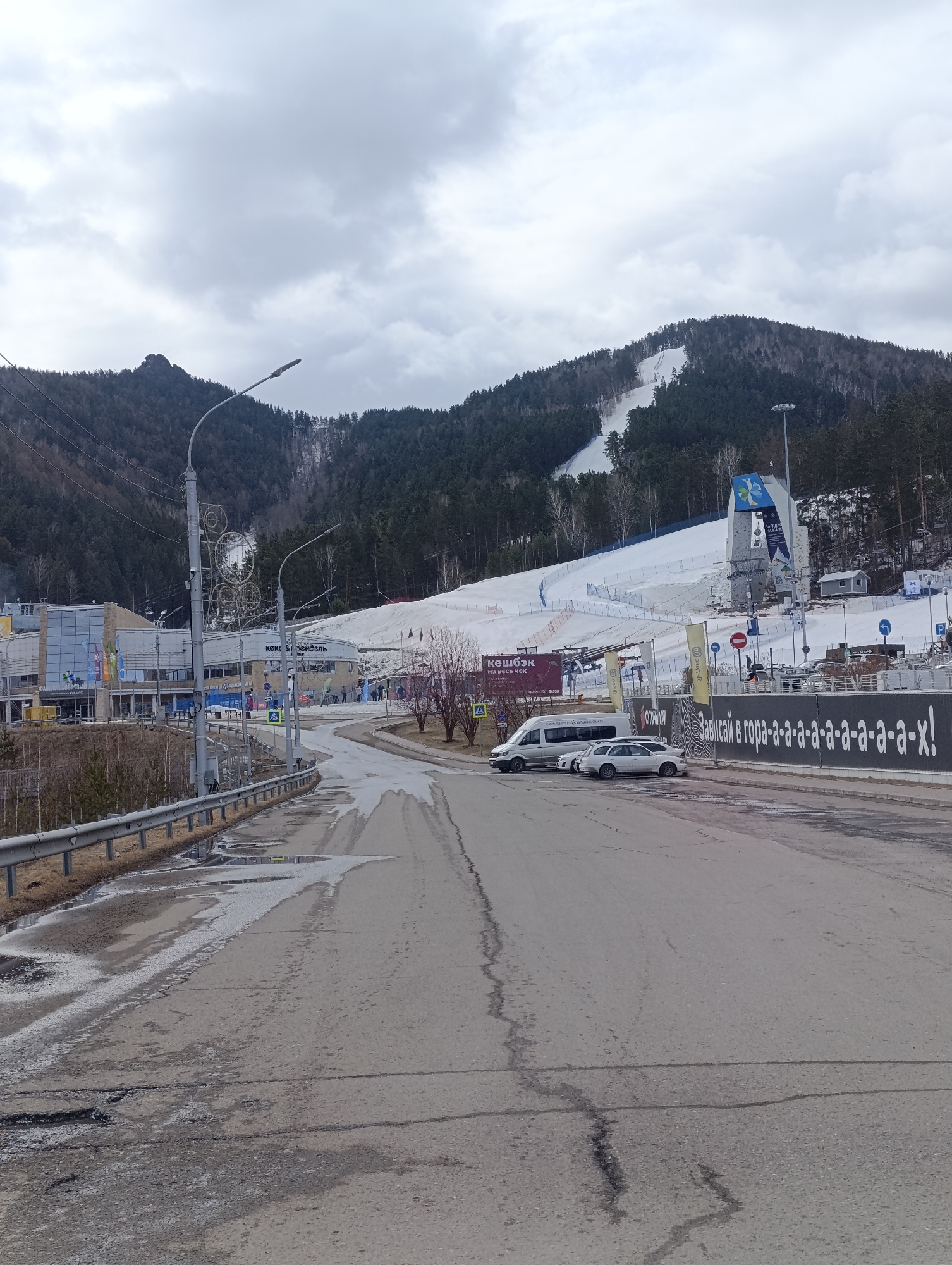 The image depicts a view of the Bobrovy Log ski resort in Krasnoyarsk, Russia, on an overcast day. The foreground is dominated by a wet and cracked asphalt road. A metal guardrail runs along the left side of the road. A white van and a white car are parked near the right side of the road. Beyond the vehicles, there's a black fence with white lettering in Russian, advertising the resort. In the background, the ski slopes are visible, with patches of snow running through the forested hillside. A building with multiple flags, ski lifts and a climbing wall structure are present near the base of the slopes. The sky is overcast.