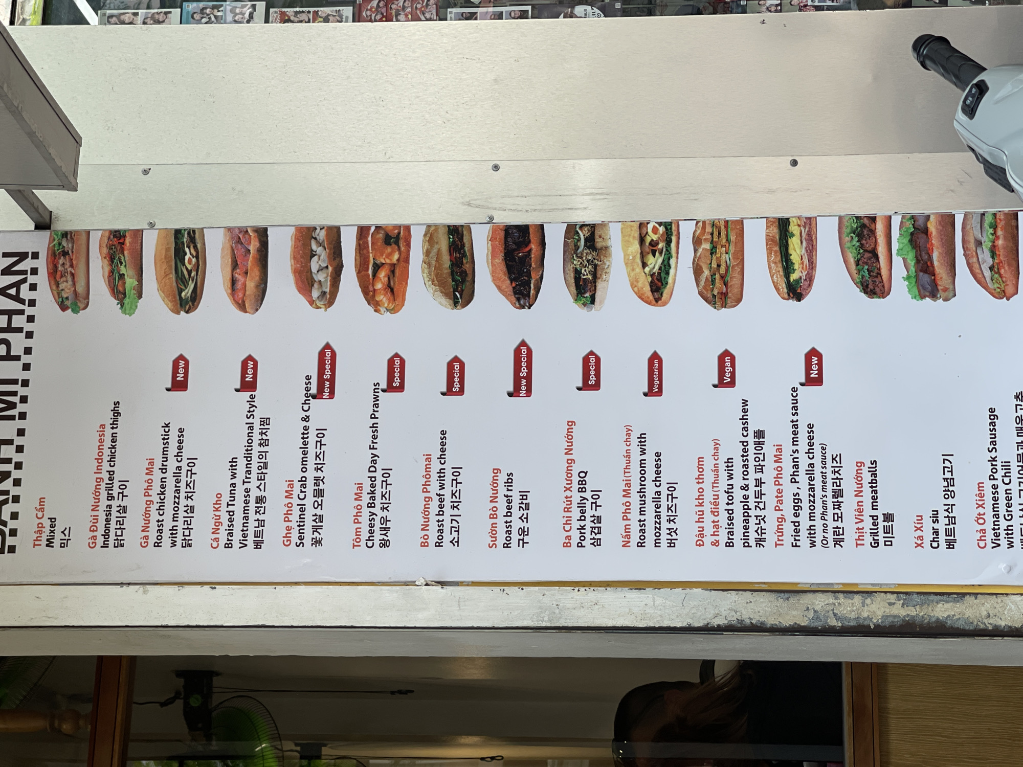The image shows a close-up of a menu board for Banh Mi Phan, a Vietnamese sandwich shop, likely located in Nha Trang, Vietnam. The menu is in Vietnamese, English, and Korean, displaying various banh mi options with corresponding photos. The menu includes descriptions of sandwiches like "Ga Đùi Nướng Indonesia" (Indonesia grilled chicken thighs), "Cá Ngừ Kho" (Braised Tuna), and vegetarian/vegan options. The board is rectangular with a white background and dark text.
