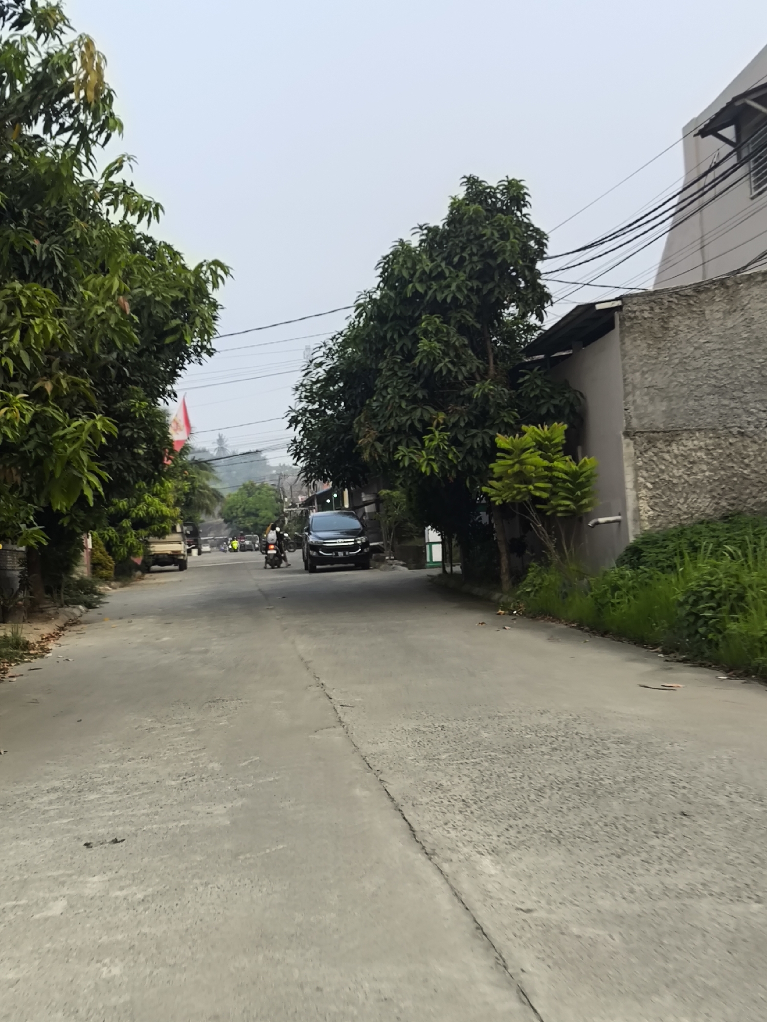The image shows a paved road in Cilegon, Indonesia, with trees lining the left side and buildings on the right. Several vehicles, including a white pickup truck, a black car, and motorcycles, are present on the road. One motorcycle has a rider, and the black car appears to be moving. A red and white flag is visible on the left side of the road, attached to a pole. The sky is overcast, and the environment appears to be a residential or mixed-use area within the city. There are electrical wires running overhead, typical of urban infrastructure. The road itself shows some signs of wear and tear, with cracks and uneven patches. The vegetation is lush and green, suggesting a tropical climate.