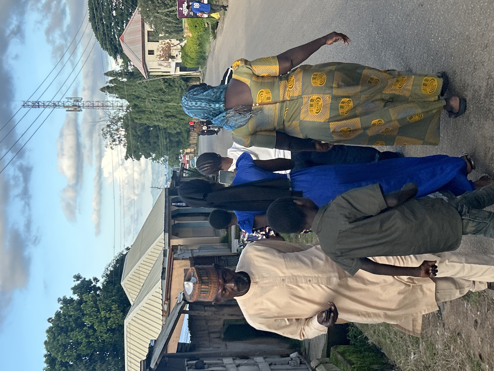 In the city of Jos, Nigeria, a group of people are walking down a paved road. The group includes an elderly man in a light-colored kaftan and a patterned cap, a boy in a dark green shirt, a woman in a blue dress, and a woman in a long, patterned green and yellow dress with a blue headscarf. Behind them, other individuals are visible, and further down the road, a person on a motorcycle is approaching. The environment consists of houses with corrugated metal roofs, trees, and a cell tower in the distance. The sky is cloudy.