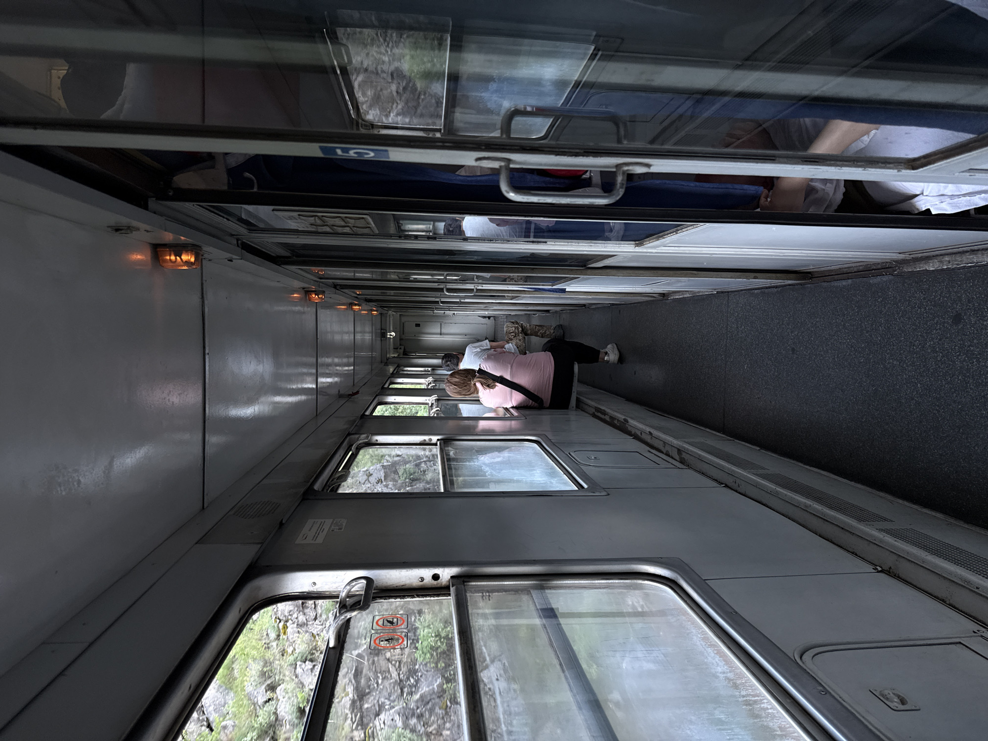 The image, taken in Klopot, Montenegro, presents the interior of a train car corridor, viewed down its length. On the right side, a sequence of large windows offers a clear view of the outside environment, characterized by rugged, rocky terrain and abundant green foliage, indicative of a mountainous region.

A woman with blonde hair, dressed in a pink top and black pants, is seated comfortably on a low surface or the floor adjacent to one of the windows, looking outwards at the passing scenery. Further down the corridor, additional passengers are visible in the distance, their forms and movements softly blurred. Reflections in the upper translucent panels of the train's interior show more passengers, one of whom appears to be wearing a face mask.

The left side of the corridor consists of what seem to be compartment doors or the solid walls of berths, fitted with metal handles. Overhead, warm orange lights are strategically placed along the ceiling, providing illumination for the passage and creating a subdued atmosphere within the train, contrasting with the natural daylight outside.

Visible text elements include the letters "LO" (or "L0") on a blue sign, seen in reflection within an upper panel. On a lower window, two distinct red circular warning symbols are present: one depicting a crossed-out human figure and another a crossed-out object, likely serving as instructional or prohibitory signs. The overall scene captures a moment of tranquil travel through a picturesque natural landscape.