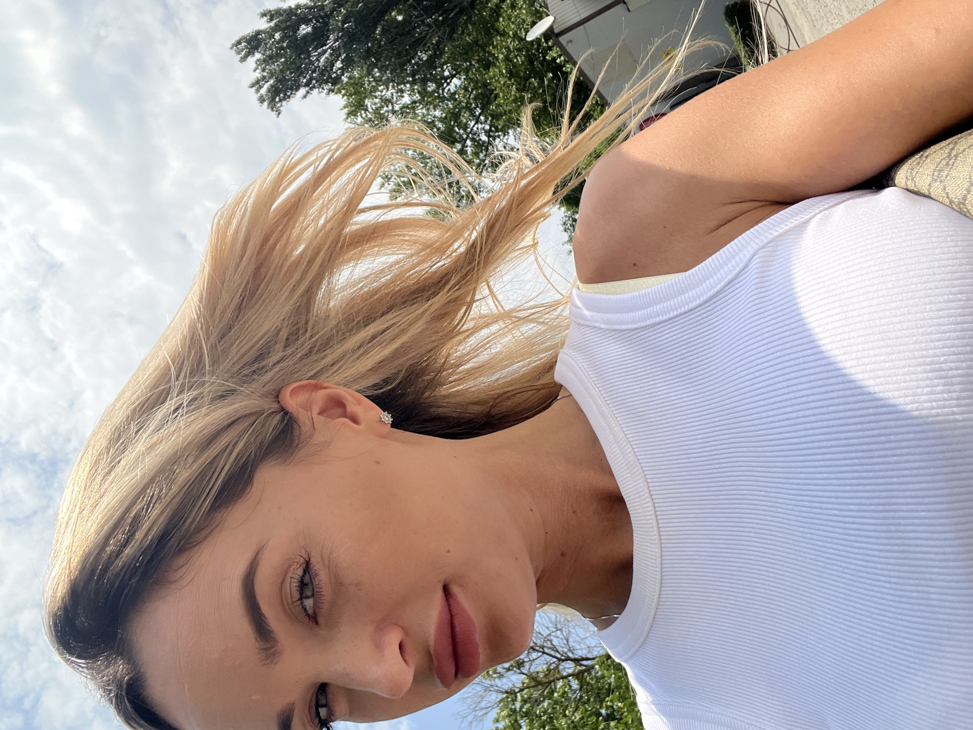 The image shows a close-up, waist-up shot of a young woman with blonde hair blowing in the wind, outdoors on a sunny day. She is looking towards the camera with a slight smile. She is wearing a white ribbed tank top, and a delicate necklace can be seen around her neck. She has a small diamond stud earring in her visible ear. The background shows a blue sky with white clouds, green trees, and what appears to be a building with a satellite dish. The image's composition suggests it was taken with a mobile phone, possibly as a selfie, given the angle and the way the woman is framed. The location is noted as Fontanka, Ukraine.