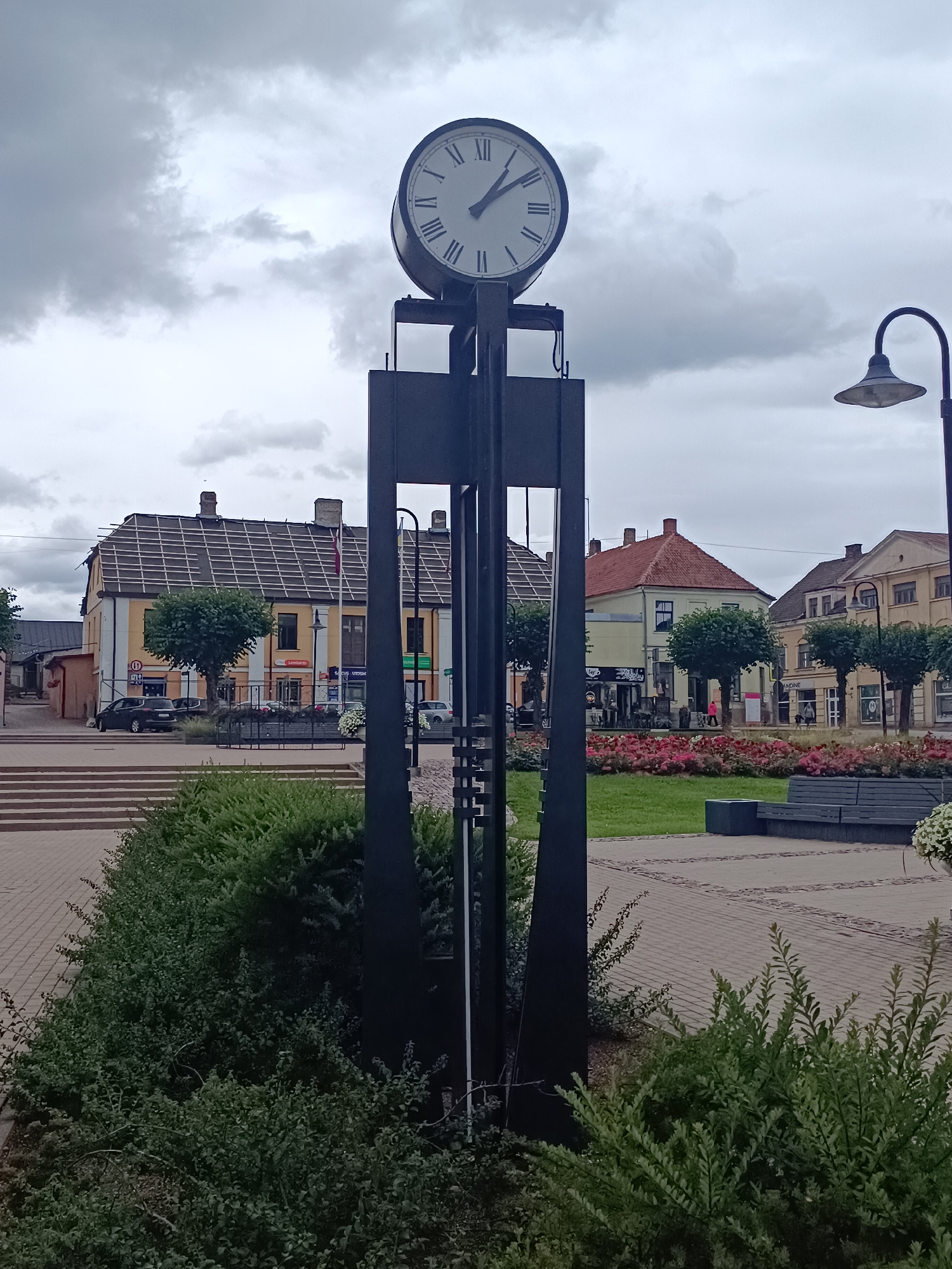 The image shows a public square in Saldus, Latvia, dominated by a tall, black, modern clock tower with a Roman numeral clock face. The clock shows approximately 10:10. In the background, there are several buildings with traditional architecture, some with shops and businesses on the ground floor. A streetlamp with a curved arm is visible to the right of the clock tower. The square features paved walkways, landscaped green areas with bushes and flowerbeds, and some benches. A few cars are parked on a street adjacent to the square. The sky is overcast with grey clouds. There are no people prominently featured in the image. The scene appears to be a quiet, daytime view of a town center.