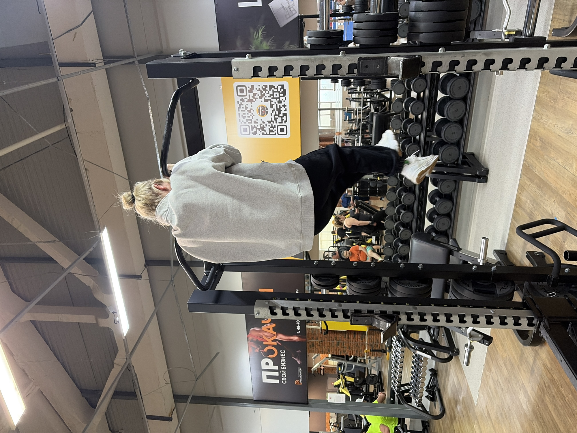 A woman in a gray sweatshirt and black pants is doing a pull-up in a gym. The gym is well-equipped with weights, dumbbells, and other exercise machines. The gym is located in Chaikovsky, Russia, as indicated by the Russian text on the advertisement. Other people are working out in the background.