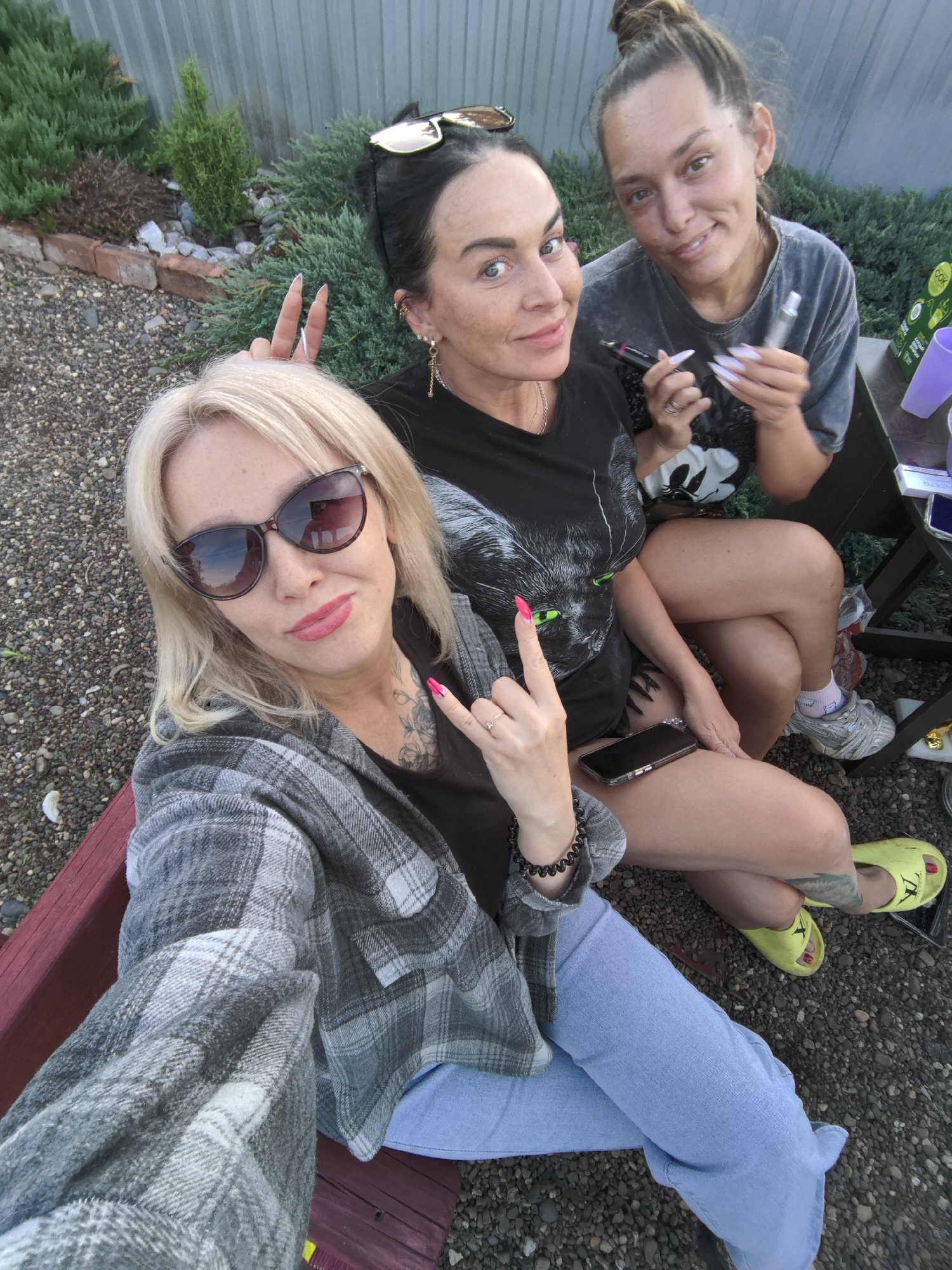 A selfie captures three women posing outdoors, likely in a backyard or garden setting. The ground is covered in gravel, and green bushes line a corrugated metal fence in the background. The lighting suggests it is late afternoon or early evening.
In the foreground, a blonde woman wearing dark sunglasses, a grey plaid shirt over a black top, and light blue jeans is holding the camera. She has bright pink lipstick and matching nail polish, with a tattoo visible on her neck and chest, and makes a "rock on" hand gesture. She is seated on a red wooden bench.
Next to her, a woman with dark hair, wearing a black t-shirt with a large cat face graphic and sunglasses pushed up on her head, is also smiling. She displays long, light-colored nails and makes a "peace" or "victory" sign with her fingers. A black smartphone rests on her lap. Neon yellow sandals are visible on the feet of the woman behind her, who is likely the middle woman's.
Further to the right, a third woman with brown hair tied in a bun is smiling directly at the camera. She wears a grey t-shirt, short shorts, white socks with a pink design, and grey sneakers. She holds two vape pens in her hands, also showcasing long, sharp, light-colored nails. On a small wooden table beside her, purple plastic cups, a small white box, and a green container with the text "COK" are visible.
All three women appear relaxed and happy, enjoying a casual social gathering outdoors.