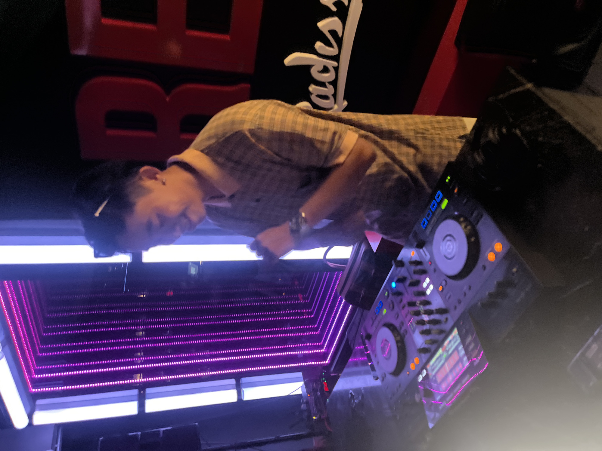 A person is seen from the side, bending over a DJ setup. They appear to be a DJ, with their hands on the equipment, adjusting the controls. The DJ is wearing a plaid shirt and has short dark hair. The DJ setup includes turntables and a mixer with various buttons and lights. The background is dark, with some red lettering visible on the upper left, which appears to say "BB". To the right of the DJ, there is white text on a black background that reads "lacks". The scene is illuminated by vibrant purple and white LED lights, creating a club-like atmosphere. The lighting is dim, suggesting it is nighttime or inside a venue. The DJ's posture and focus suggest they are engaged in mixing music. The overall impression is that of a DJ performing in a music venue or club. The image is oriented sideways.