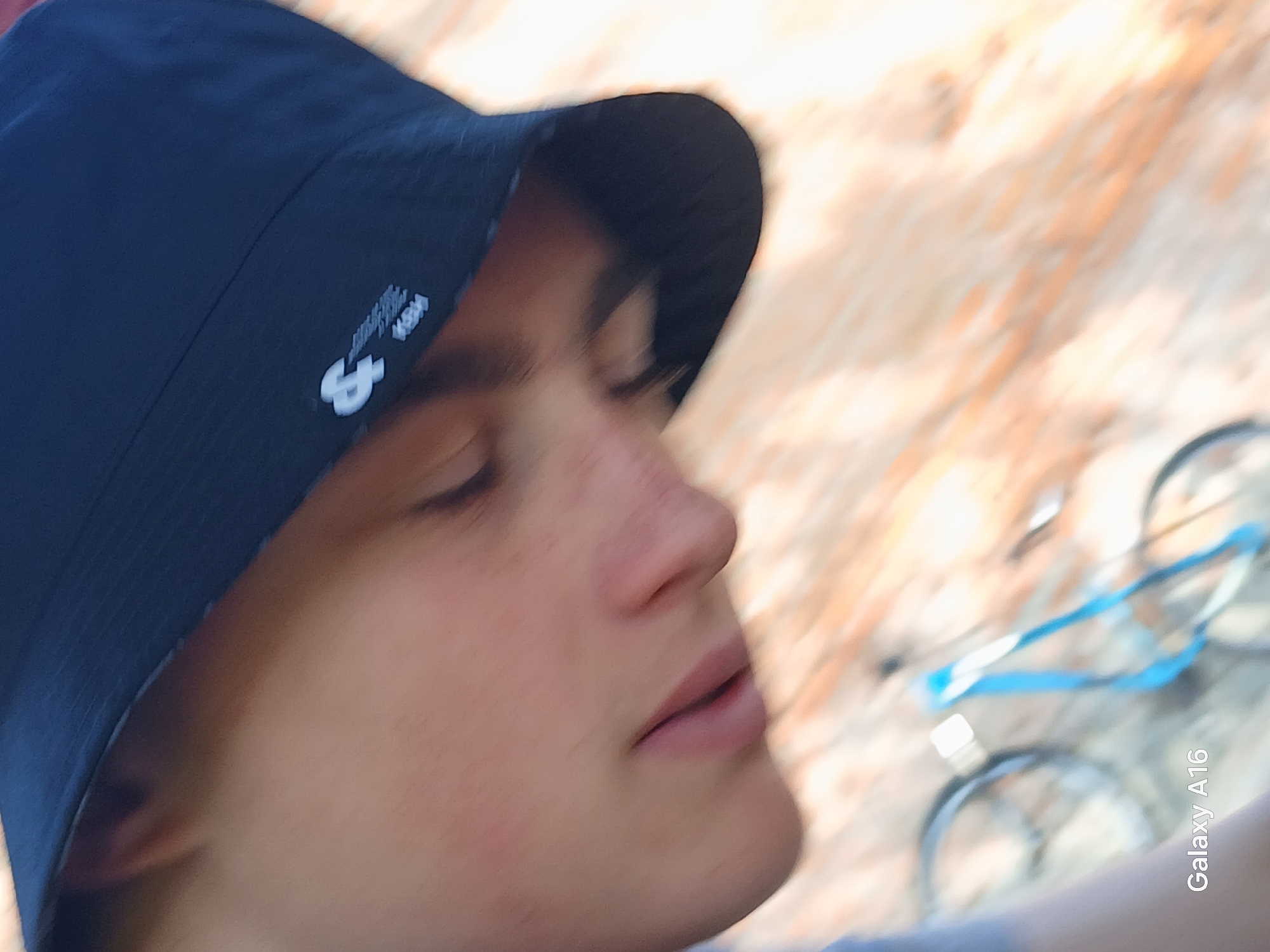 The image is a close-up, angled shot of a person wearing a dark bucket hat. The person's face, partially obscured by the hat, is tilted downwards, with their eyes closed or looking down. The skin appears to have light freckles. The hat has a white logo on the side. In the blurred background, a bicycle with a blue ribbon or strap is visible. The scene appears to be outdoors, with a wooden surface beneath, suggesting a park, a yard, or a wooden deck. The bottom left corner of the image shows the text "Galaxy A16," indicating the device used to capture the photo. There is no specific information within the image that places it in Ustyvytsia, Ukraine.