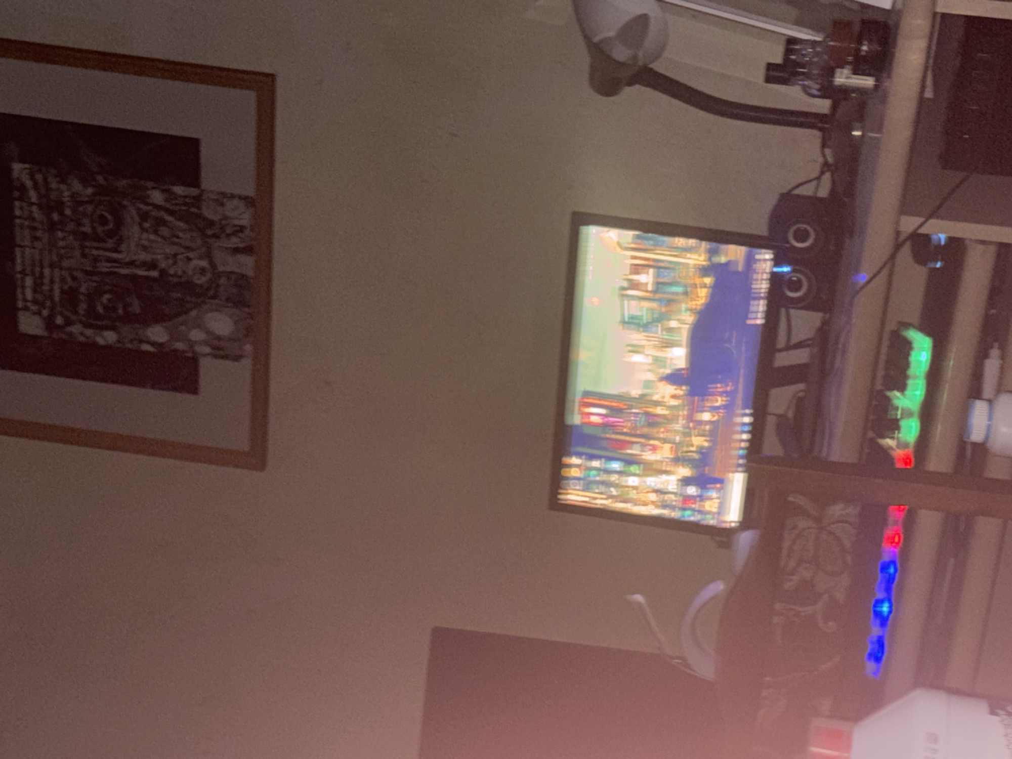 The image shows a partially illuminated room with a framed artwork on the left wall. A computer monitor displays a vibrant image of an urban scene, possibly depicting the city of Yerevan, Armenia. The desk arrangement includes speakers, computer components with green and red LED lights, and other unidentified items. The scene is dimly lit, creating a somewhat hazy atmosphere.