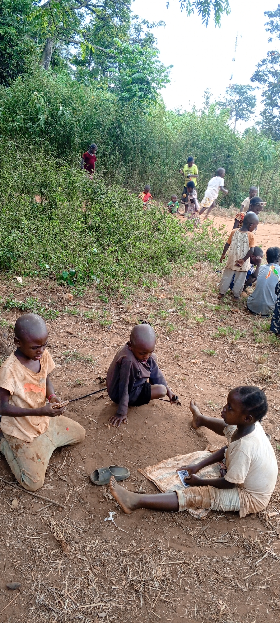 The scene is an outdoor clearing in Ngalula, Democratic Republic of the Congo. In the foreground, three children sit on the ground, engaged in what appears to be a game with playing cards. One child sits cross-legged, holding cards; another is focused on a small object, possibly a toy; and a third is seated on a cloth, observing the others. To the left, there is a single sandal. In the background, other children are either standing, running, or playing near a thick line of vegetation. The ground is dirt with sparse patches of grass. Trees are visible in the distance. The clothing worn by the children is worn and soiled.