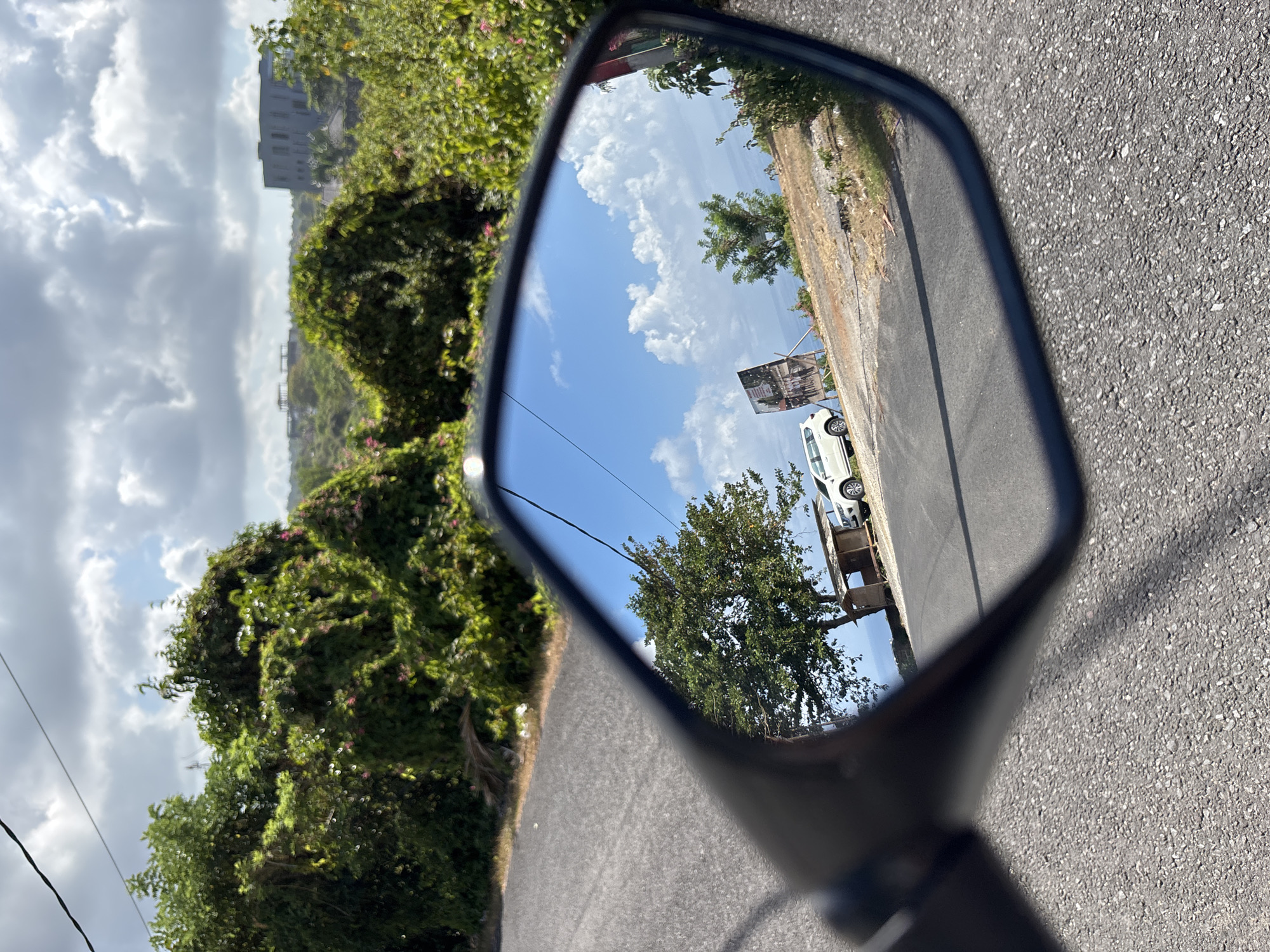 The image captures an outdoor scene from the perspective of a motorcycle, prominently featuring its rearview mirror. The overall view suggests a daytime setting on a road in a tropical environment, consistent with Kuta Selatan, Indonesia.
Inside the rearview mirror, a clear blue sky with scattered white, fluffy clouds dominates the upper half of the reflection, indicating sunny weather. A paved road is visible, with a white car parked on its right shoulder. To the left of the road within the mirror, a large green tree stands, while further back on the right, a billboard with an indiscernible image or text is partially visible behind the parked car. The scene reflected in the mirror is brightly illuminated by direct sunlight.
Outside the mirror's frame, the immediate foreground consists of a textured asphalt road surface, leading towards the left. Lush, dense green foliage, possibly vines or bushes with hints of pink flowers, lines the left side of the road and extends into the background. Further in the distance on the left, a multi-story building with a light facade is visible. The sky outside the mirror appears partly cloudy and slightly less bright than the sun-drenched reflection. Thin power lines are also visible stretching across the lower-left portion of the overall image. No specific activity or emotion is discernible beyond the static road scene.
