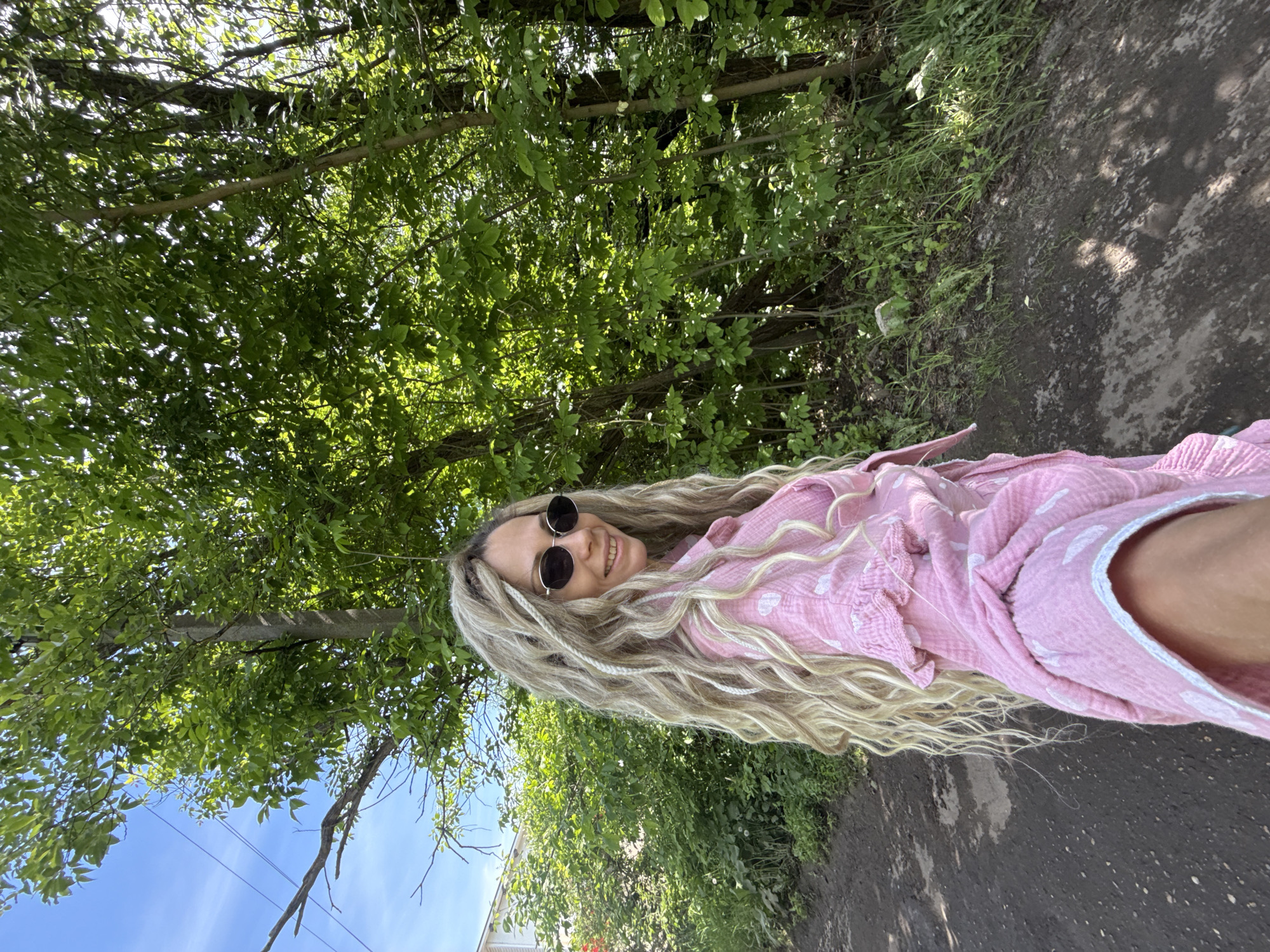 A woman with long, wavy blonde hair, featuring lighter braided sections, is captured in a close-up, selfie-style photograph in Rusanovo, Russia. She wears dark, round sunglasses and smiles directly at the viewer. Her attire consists of a light pink, long-sleeved button-up shirt with white heart-shaped patterns and ruffled detailing near the shoulders. The setting is outdoors, on a dirt path with visible soil variations, possibly indicative of dampness, extending into the foreground. To the woman's left, green shrubs and undergrowth lead to a distant light-colored building under a clear blue sky with overhead utility lines. To her right and in the background, a dense expanse of vivid green foliage and tree trunks creates a wooded environment, with dappled sunlight filtering through the leaves, casting shadows on the ground.