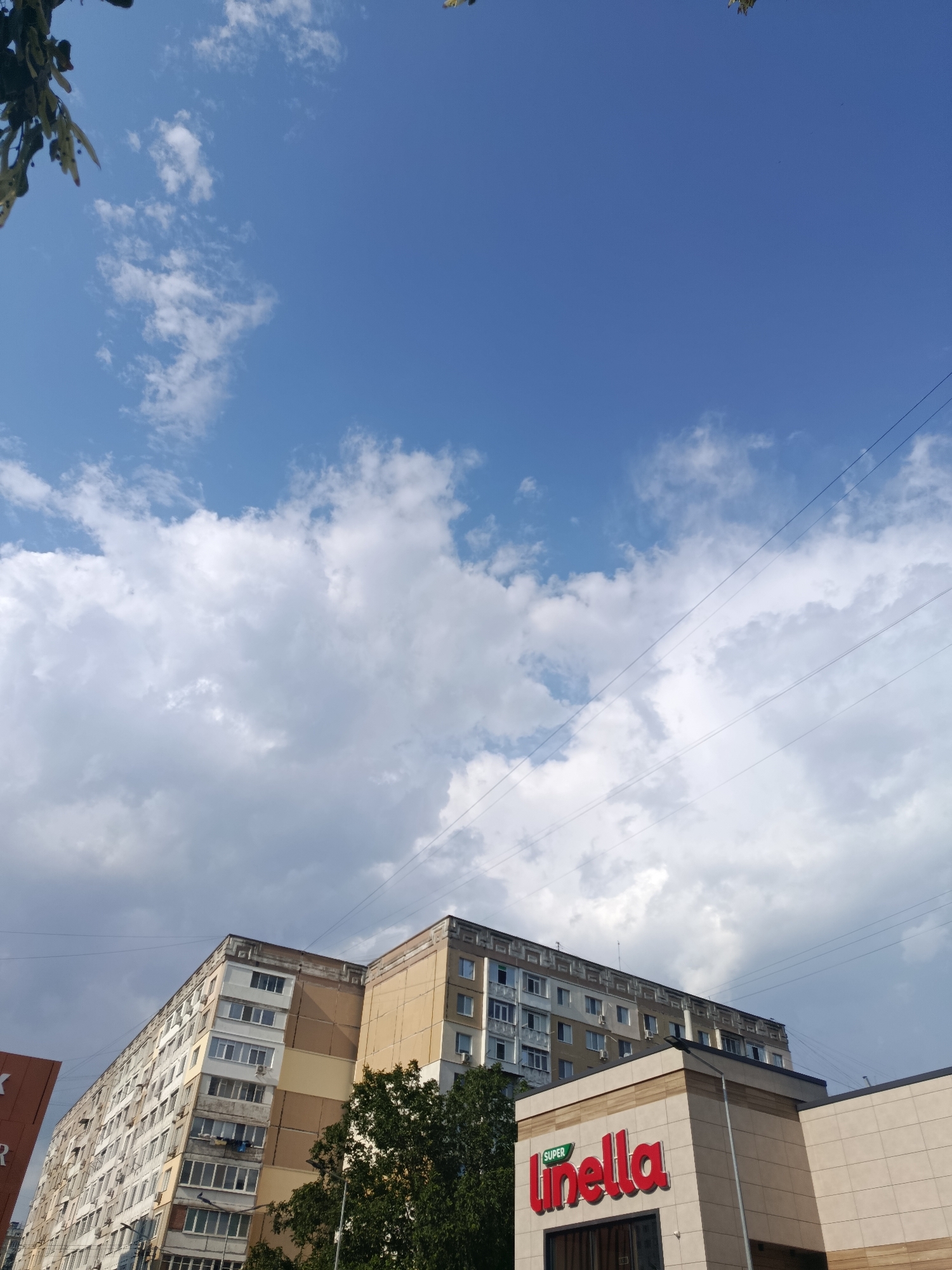 The image depicts a view of apartment buildings and a commercial establishment in Chișinău, Moldova. In the foreground, a modern building with a beige facade features prominent red lettering that reads "SUPER linella." Behind this building, a tall, older apartment complex with multiple floors and numerous windows is visible, showing signs of wear. To the left, part of another sign with orange and white lettering is partially visible. Lush green trees are interspersed between the buildings. The sky above is a bright blue, dotted with fluffy white clouds. Electrical wires crisscross the sky, strung between unseen poles. The perspective suggests an outdoor, daytime scene in an urban environment.