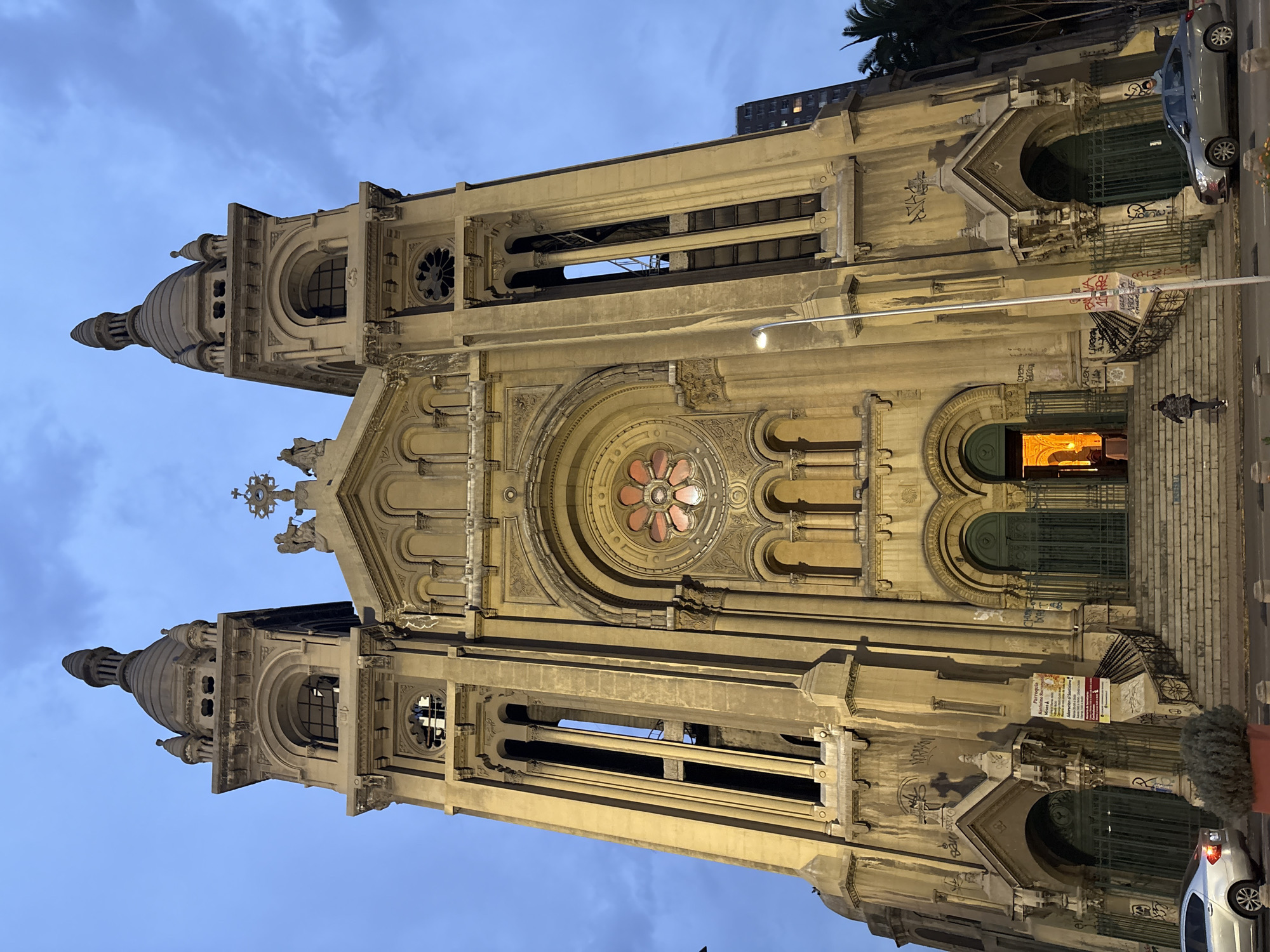 The image depicts the Basilica of the Sacred Heart of Jesus in Santiago, Chile, at dusk. The imposing neoclassical facade of the church is illuminated by interior lights, casting a warm glow. The sky is overcast with dramatic clouds. Two bell towers flank the central structure, each topped with a spire. Arched windows and intricate architectural details are visible on the facade. A circular rose window with stained glass is centrally located above the entrance. Steps lead up to the main entrance, where two green metal gates are open. A person is seen walking up the stairs. In the foreground, a street with cars and a streetlight is visible. Graffiti is present on the lower sections of the church's facade. Lush palm trees can be seen to the right of the building.