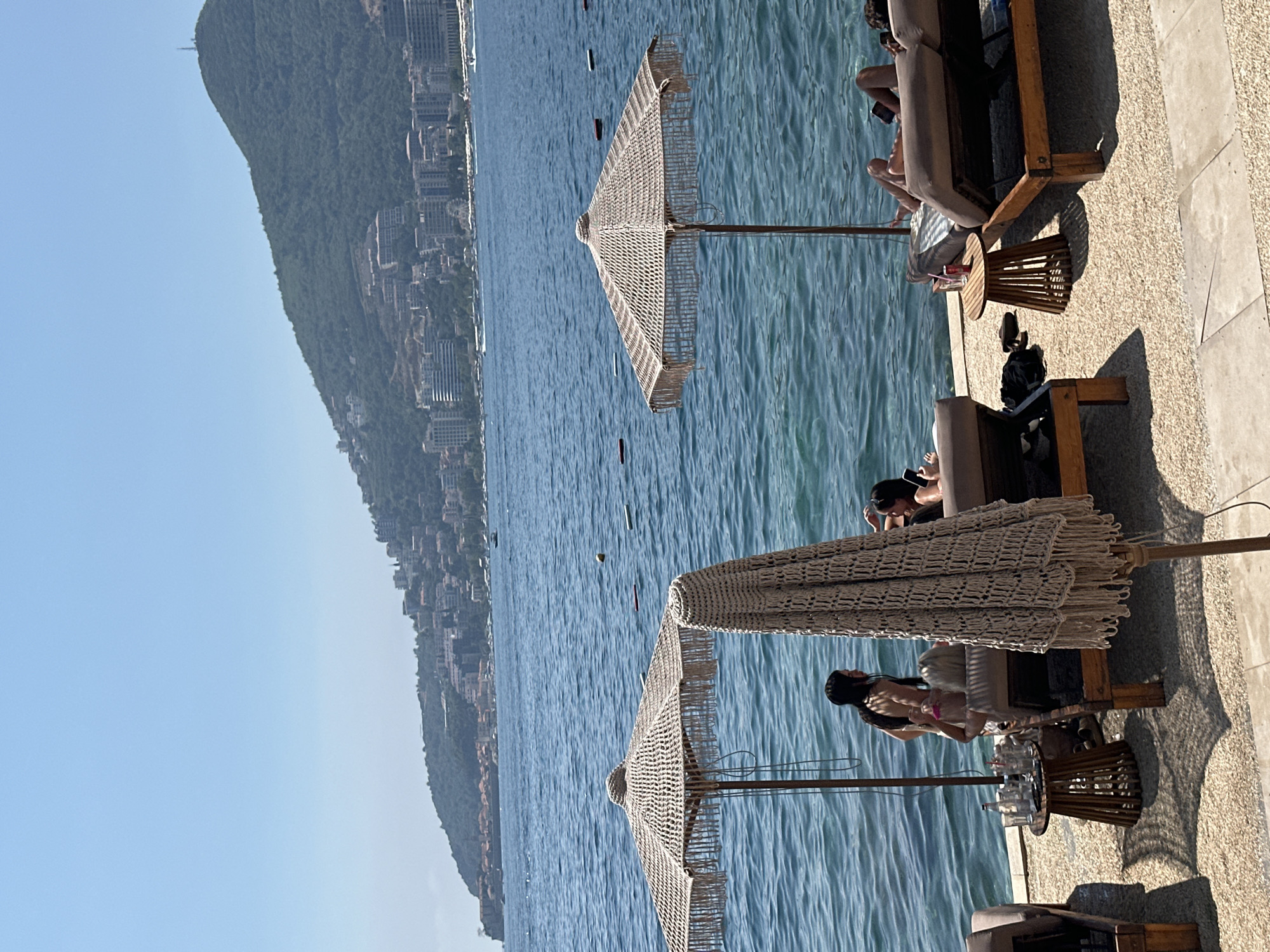 In Budva, Montenegro, on a sunny day, a beach scene unfolds with a clear blue sky and calm turquoise waters. In the foreground, two macrame beach umbrellas stand, one open and the other folded. Beneath the open umbrella, two individuals recline on lounge chairs, one with a book. Beside them, a small wooden side table holds drinks. To the left, another lounge chair is occupied by a person with a phone, and a side table displays more drinks and a bottle. Closer to the water's edge, two women are in the water, one standing and the other sitting. Further back, on the right side, two more people are relaxing on lounge chairs, one with a book and the other looking at a phone. The background features a lush green mountain covered in buildings, suggesting a cityscape overlooking the bay, with several boats dotting the water.