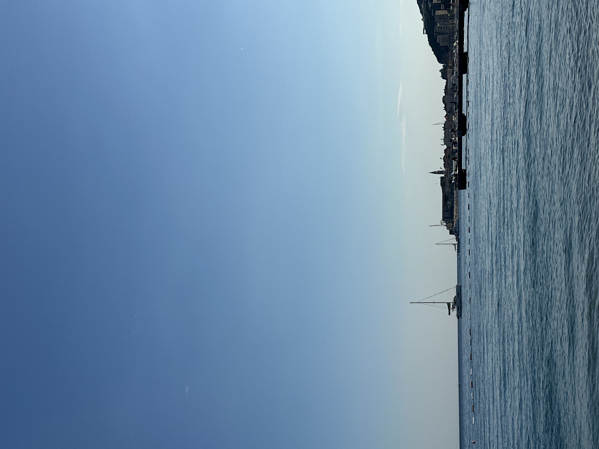 The image captures a view of Budva, Montenegro, from the sea. The sky is a gradient of blue, transitioning from a darker hue on the left to a lighter shade on the right. The Adriatic Sea is on the right, showing small waves. Several sailboats are visible on the water, indicating maritime activity. The coastline of Budva is visible in the background, with buildings clustered along the shore and extending up the hillside, suggesting a densely populated area. The overall atmosphere is bright and clear, likely a sunny day. A line of buoys runs perpendicular to the horizon.
