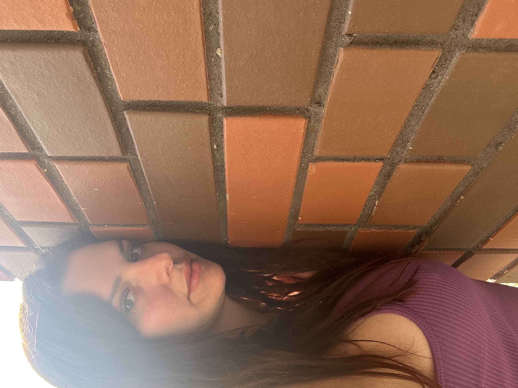 A woman leans her head against a brick wall. She has long, dark hair and is wearing a purple tank top. The wall is made of reddish-brown bricks of varying shades and textures. The lighting suggests an outdoor environment.