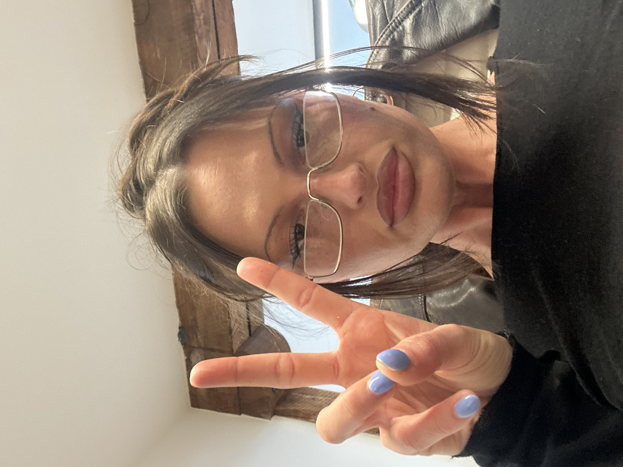 A woman poses indoors for a selfie, oriented with the camera above her. She is Caucasian, with brown hair pulled back, and wears glasses with gold frames. Her lips are full, and she is making a peace sign with her left hand, displaying light purple nail polish. She wears a black top, partly obscured by a leather jacket draped over her shoulder. The background includes a white wall, and a partially visible wooden beam, and a glimpse of a window. There is no indication of her location being in London or the United Kingdom.