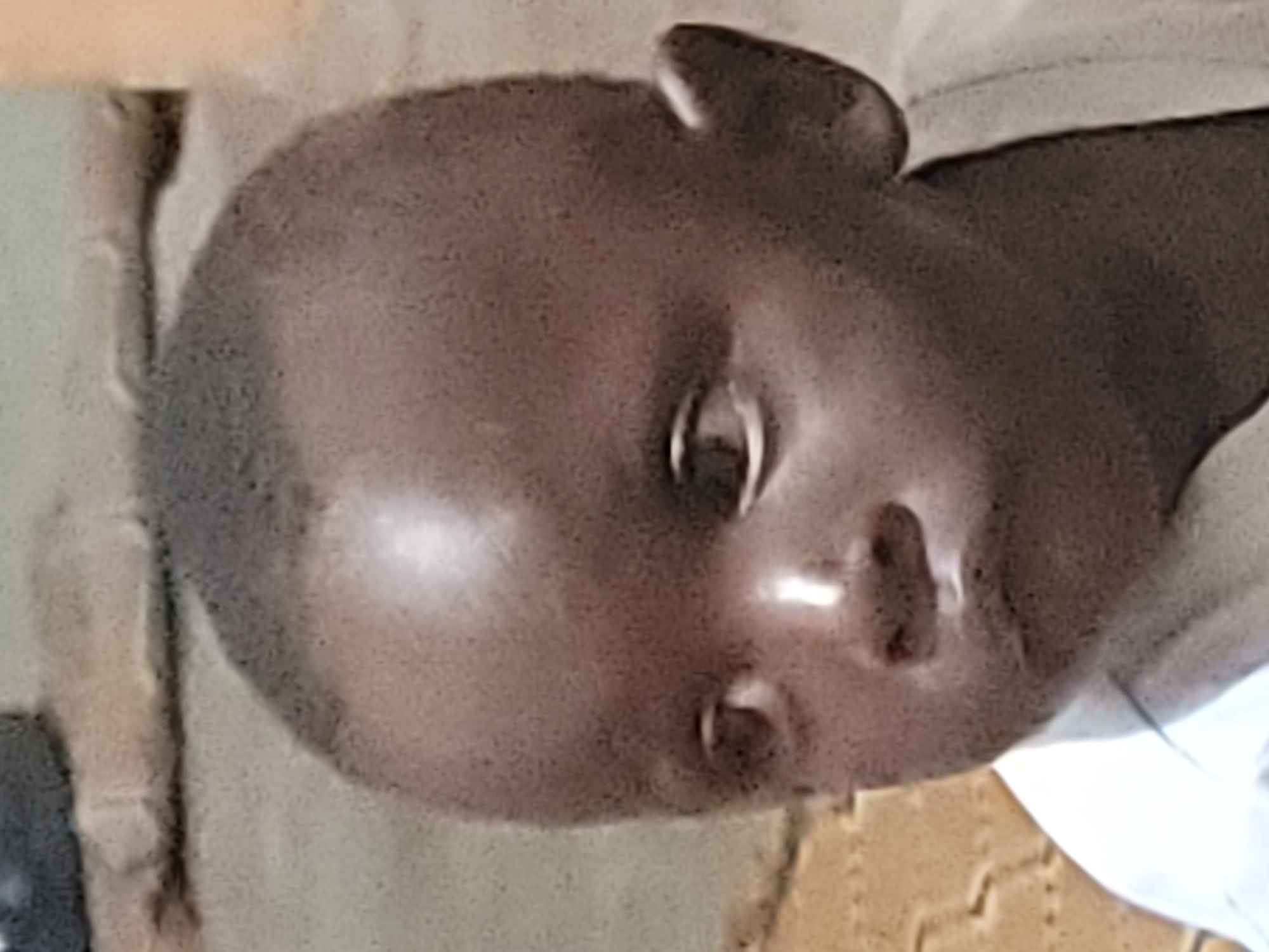 This is a close-up, head-and-shoulders, eye-level, medium shot of a young child, likely a toddler. The child has dark skin and a shaved head. The child's eyes are looking to the left of the frame, and their mouth is slightly open, forming a pout. The child is wearing a white shirt. The background is out of focus and appears to be an indoor or outdoor setting with textured surfaces. The lighting suggests an ambient light source, possibly daylight, causing a bright highlight on the child's forehead and cheek. There is no discernible specific location within Barakin Akawo, Nigeria, or any environmental context that would allow for precise localization within the city.