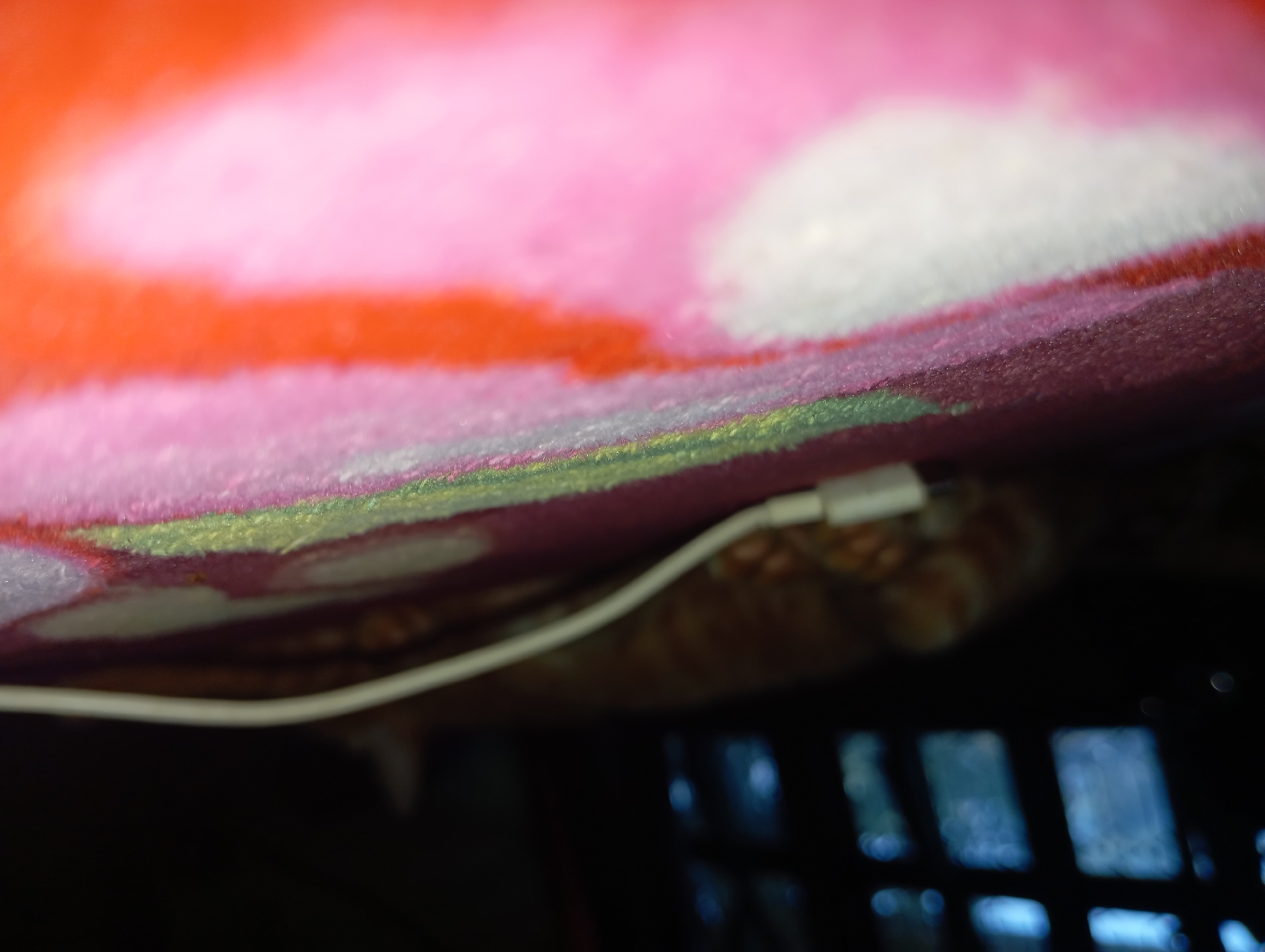 In an indoor setting, possibly a room in Rejosari, Indonesia, an out-of-focus image captures a close-up view of a white cable, possibly a charger, lying on a brightly colored, plush surface. The surface appears to be a blanket or rug with distinct sections of pink, orange, and white, interspersed with hints of green and yellow. In the background, the blurry forms of a dark grid-like structure and a partially visible orange-colored furry object, potentially a pet, are discernible. The lighting is dim, with some blueish light emanating from behind the grid structure.