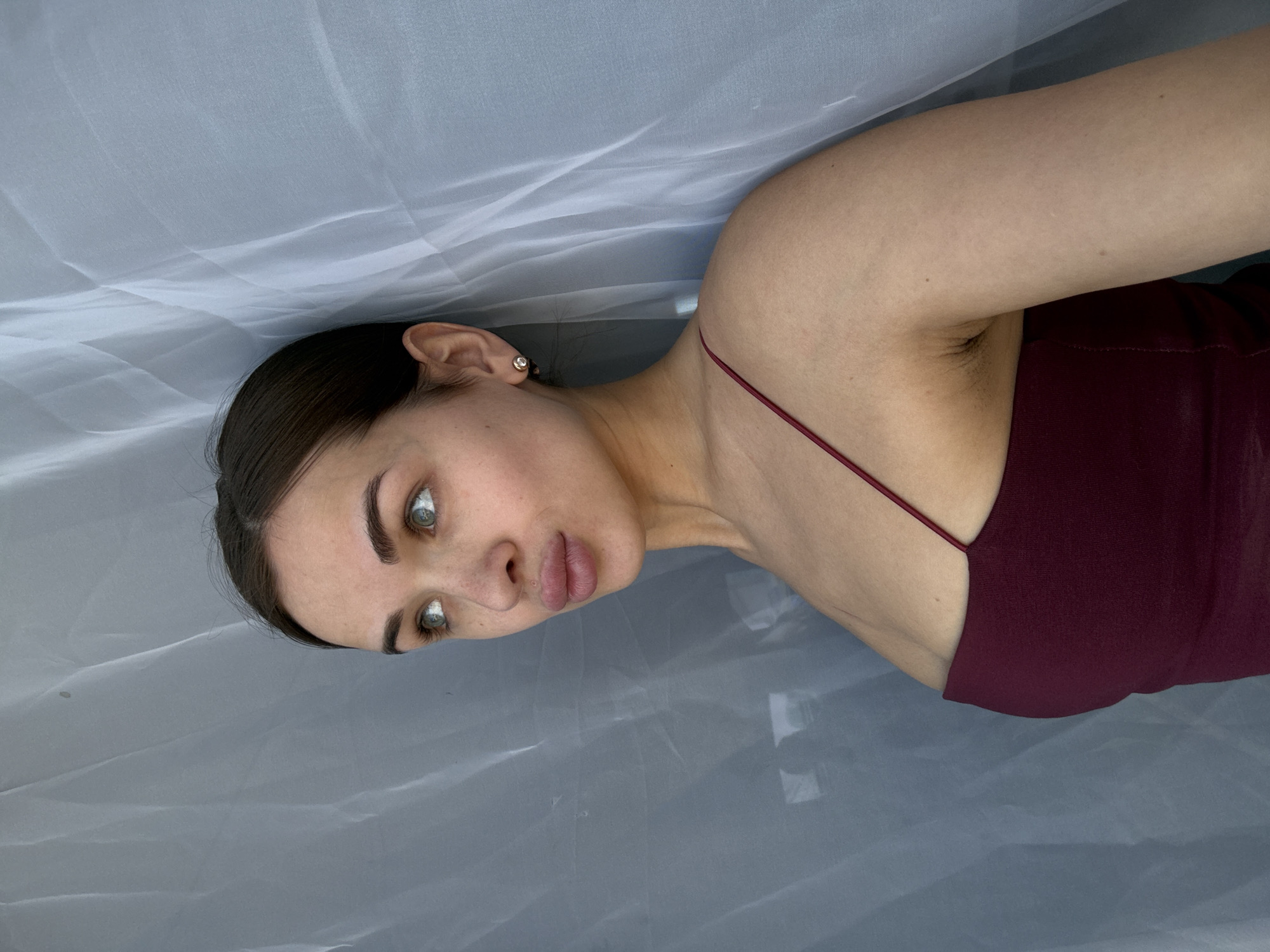A young woman lies on a grey surface, likely fabric, wearing a maroon spaghetti-strap top. She has fair skin, dark hair pulled back, and light-colored eyes. Her left ear is visible with a stud earring. The image does not contain sufficient information to identify the location as Лобаново, Russia, or to describe any actions or interactions.
