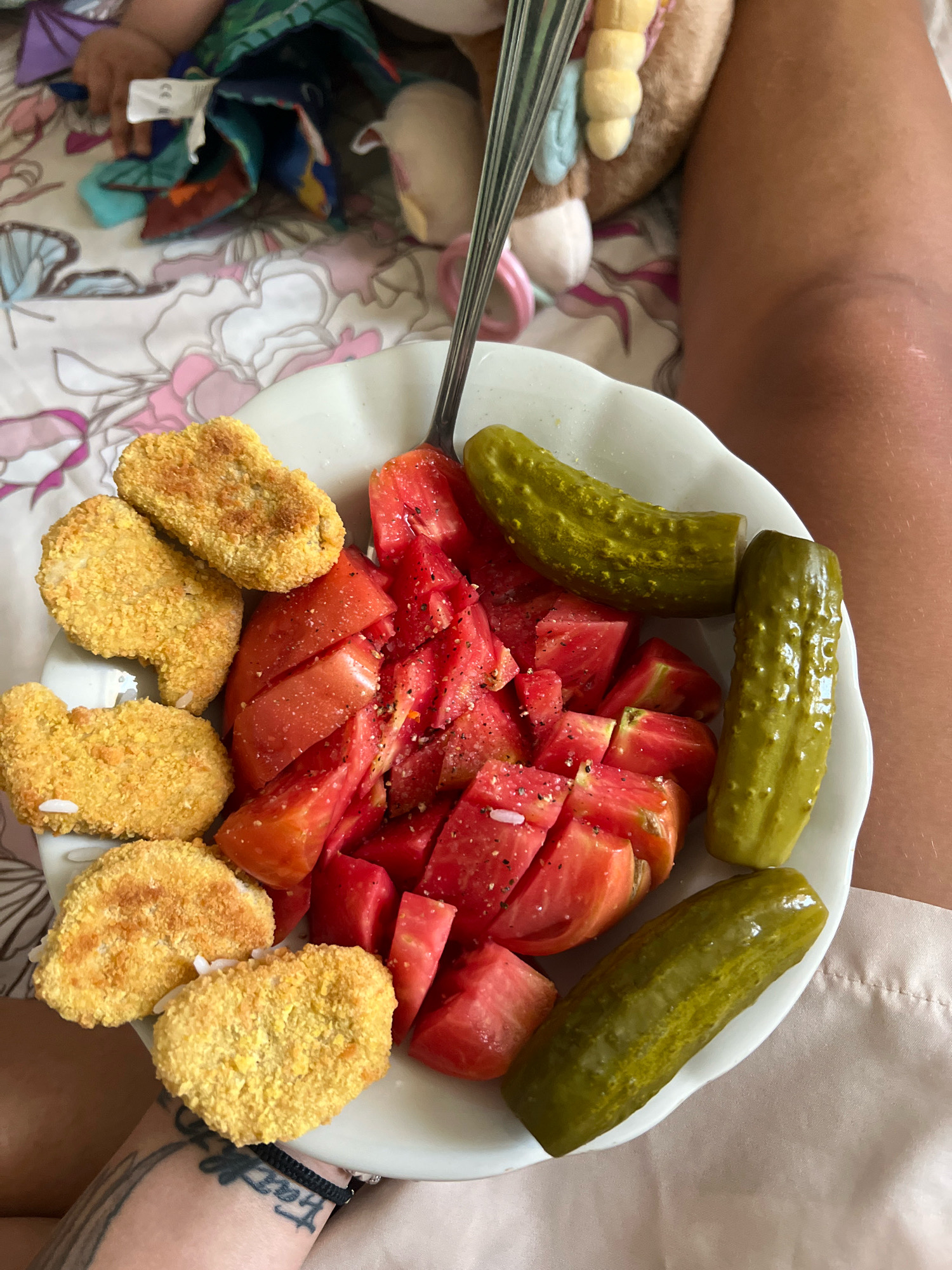 The image shows a white bowl filled with food, placed on a patterned fabric surface. The bowl contains chopped tomatoes seasoned with black pepper and salt, several dill pickles, and a cluster of golden-brown chicken nuggets. A fork is partially visible in the upper portion of the bowl. To the right of the bowl, a person's bare leg is visible, wearing a light-colored garment. In the background, a portion of a stuffed toy and a child's hand are also visible, suggesting a domestic setting. The scene does not provide any specific information that would allow for its location to be identified in the city of Plovdiv, Bulgaria.