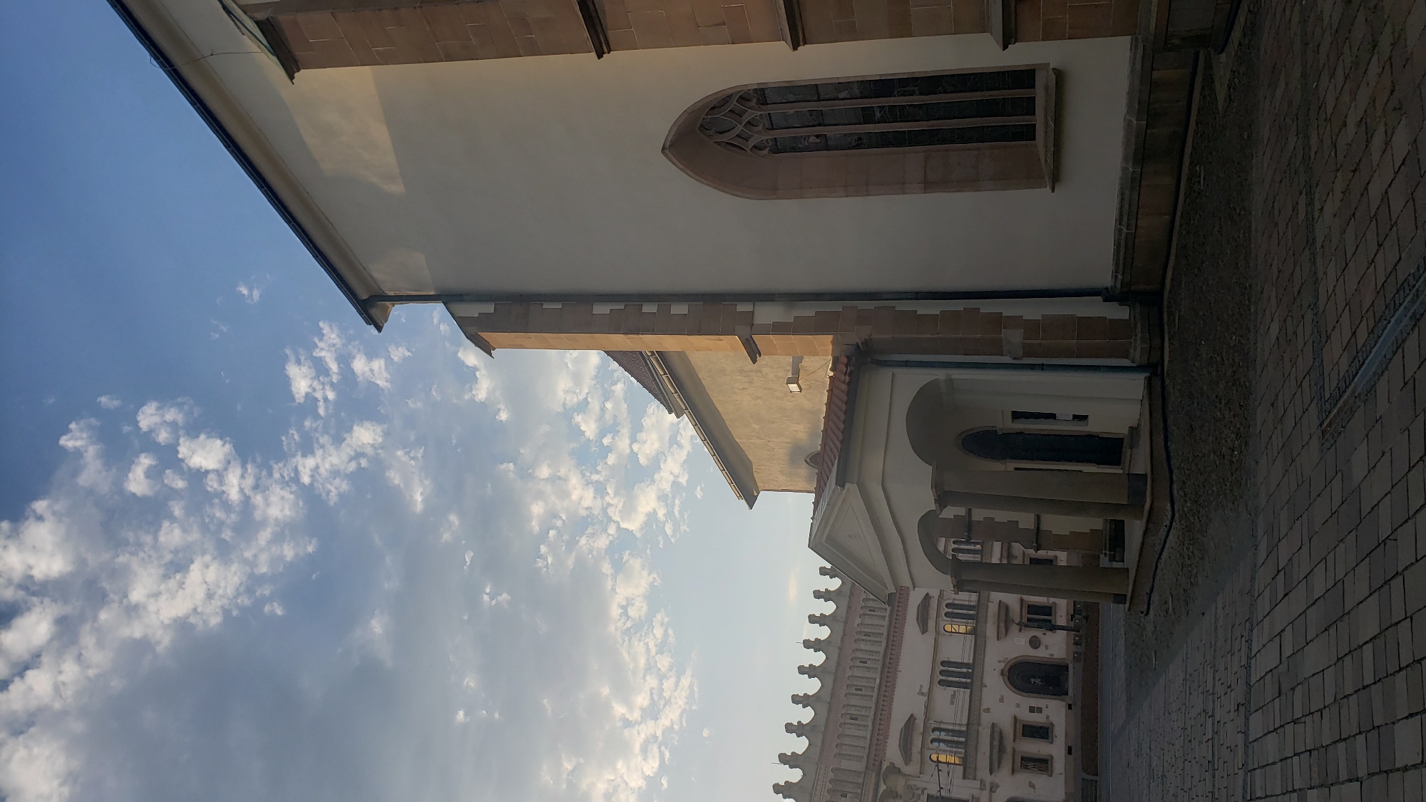 The image captures a view of St. Nicholas Cathedral in Prešov, Slovakia. The cathedral, a prominent architectural landmark, is depicted in the foreground with its white facade and arched windows. The sky above is partly cloudy, with a mix of blue sky and white clouds, indicating fair weather.
To the left of the cathedral, a street paved with cobblestones leads towards other buildings. One building visible in the mid-ground features a distinct facade with decorative elements and arched windows, suggesting historical significance and potentially a cultural or administrative function within the city. The architectural styles of the buildings suggest a European urban setting.
The scene is devoid of any visible people, emphasizing the architectural and environmental elements of the location. The lighting suggests it might be morning or late afternoon, given the shadows cast and the warm illumination on the buildings. The overall impression is one of a quiet, historic city square.