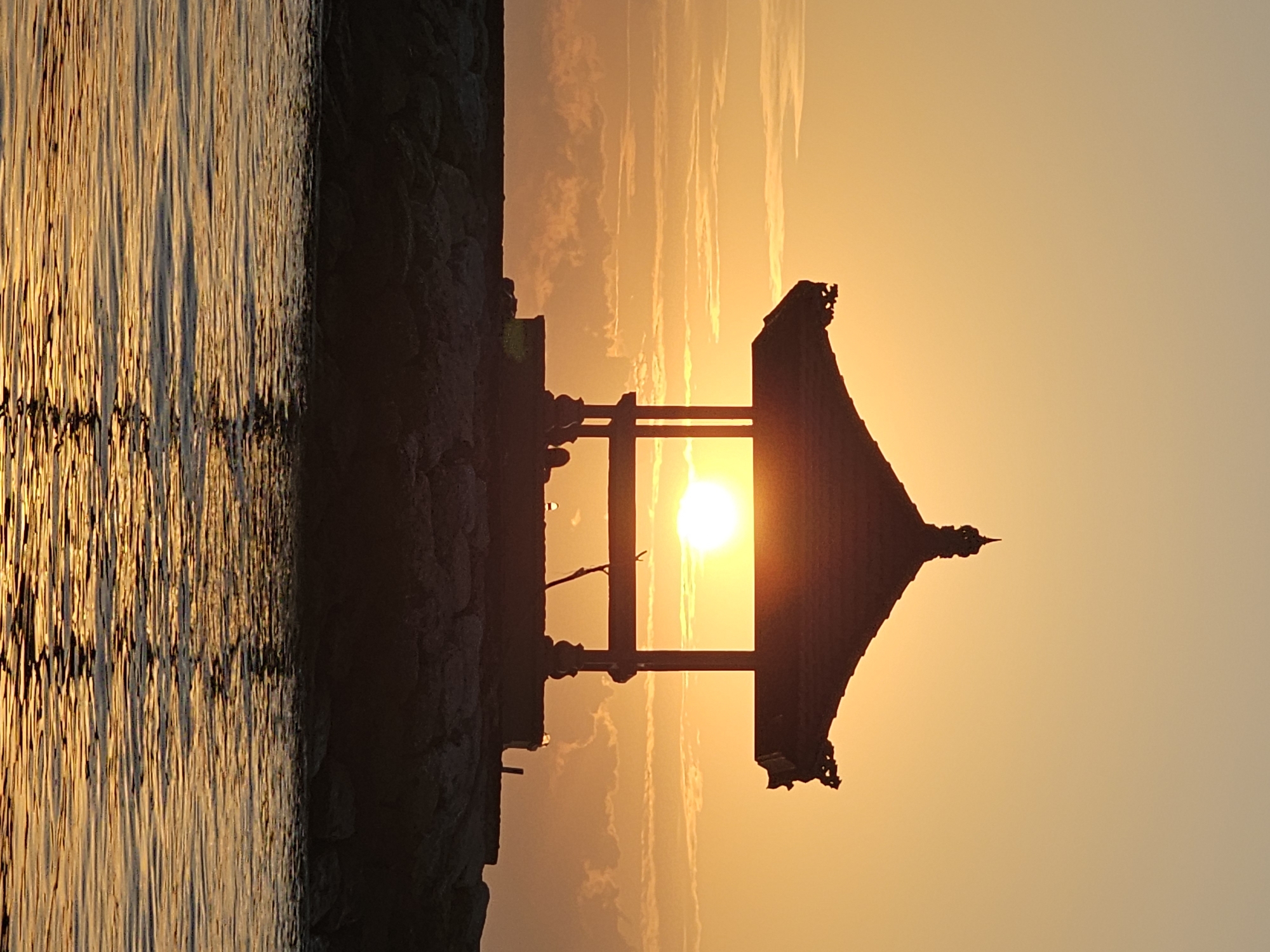 This image captures a serene sunset scene at the water's edge in Denpasar, Indonesia. A silhouette of a traditional Balinese gazebo stands prominently on a rocky embankment, framing the setting sun. The sun, a bright orb, is positioned directly behind the gazebo, casting a warm golden light across the sky and reflecting on the rippling water in the foreground. The sky is filled with soft, wispy clouds, painted in hues of orange and yellow. The gazebo itself has a distinctive tiered roof with intricate carvings. The rocky shore appears to be a breakwater, separating the calm sea from the land. The water is a deep blue with shimmering golden reflections of the sunset. There are no visible people in the image, but the silhouette suggests there may be a few figures inside or near the gazebo. The overall atmosphere is peaceful and tranquil.