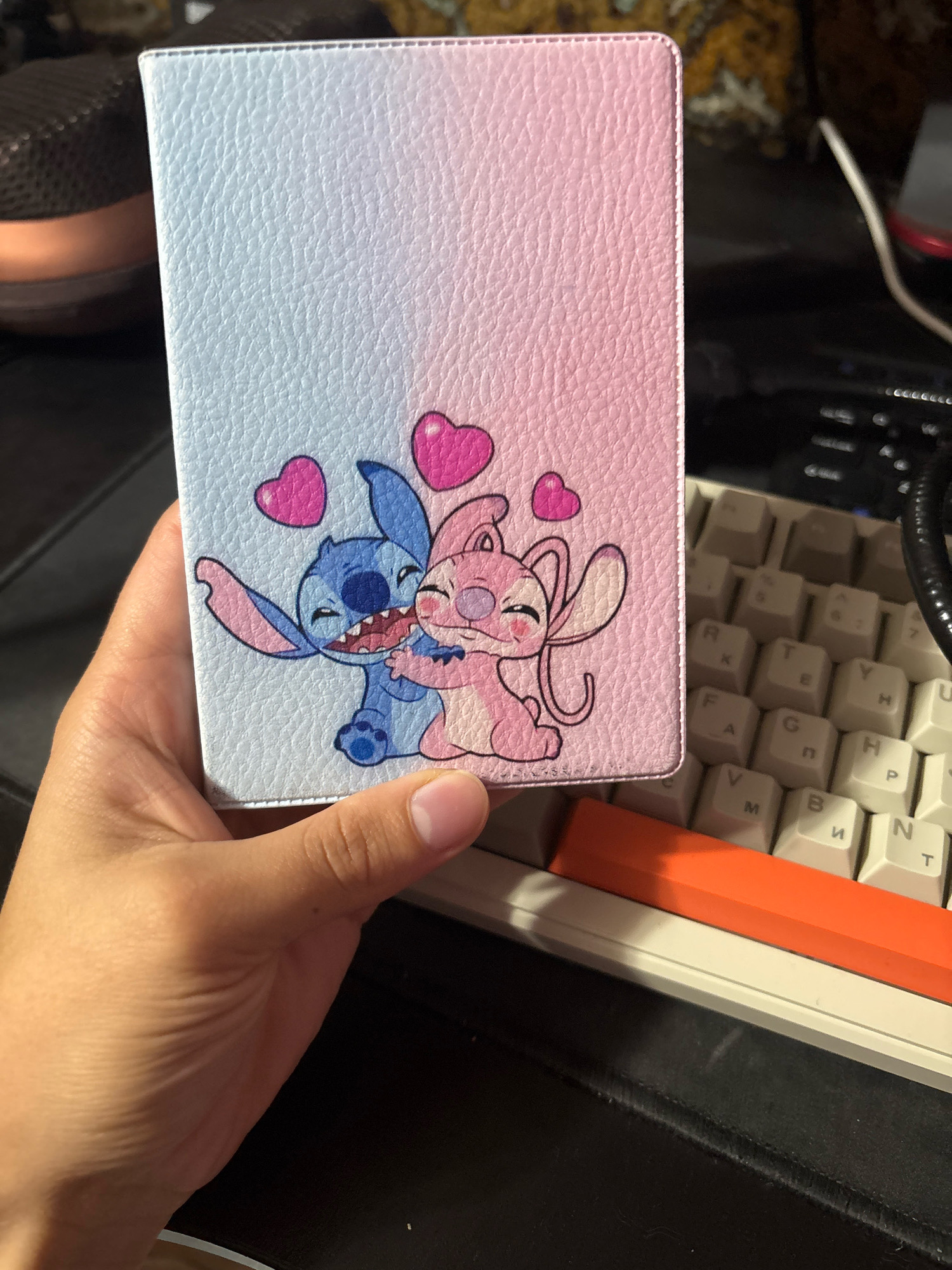 A hand is holding a light blue and pink gradient phone case with Stitch and Angel characters embracing, with pink hearts above them. The phone case is textured and has visible stitching along the top edge. In the background, a computer keyboard with beige keys is partially visible, along with a black surface that appears to be a desk or mat, and a glimpse of a dark, blurry background. An orange object is resting on top of the keyboard. The overall scene is an indoor setting, likely a workspace. The location of Novocheboksarsk, Russia, cannot be determined from the visual information provided in the image.