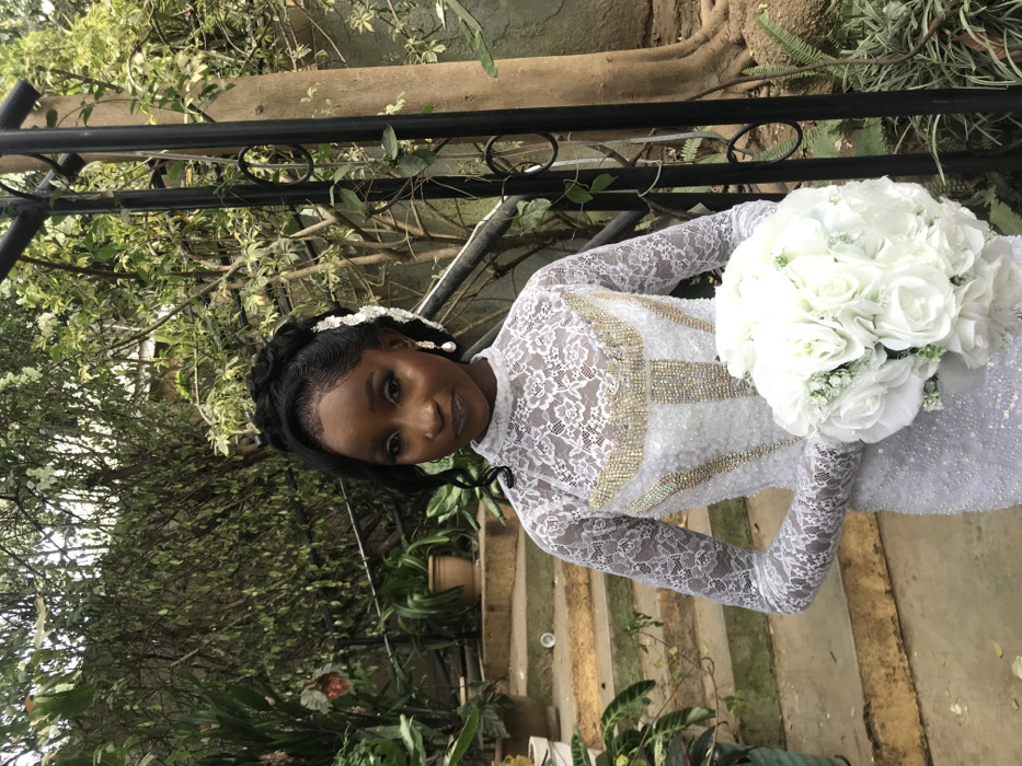 A female individual stands outdoors in what appears to be a garden or courtyard in Jos, Nigeria. She faces the viewer directly with a neutral expression. Her dark hair is styled in an intricate updo with braids and curls, accented by white floral embellishments on her left side. She wears full facial makeup, including defined eyebrows, eye makeup, and lip gloss, along with pearl drop earrings. Her attire is a white, long-sleeved, high-neck gown. The sleeves and upper bodice are made of white lace, while the mid-bodice features vertical and curved sections embellished with gold-colored rhinestones over a sequined fabric. She holds a large, spherical bouquet composed entirely of white roses in front of her. The background includes a set of ascending, weathered concrete steps with potted plants on the left, and dense green foliage, trees, and a dark, ornate metal railing on the right.
