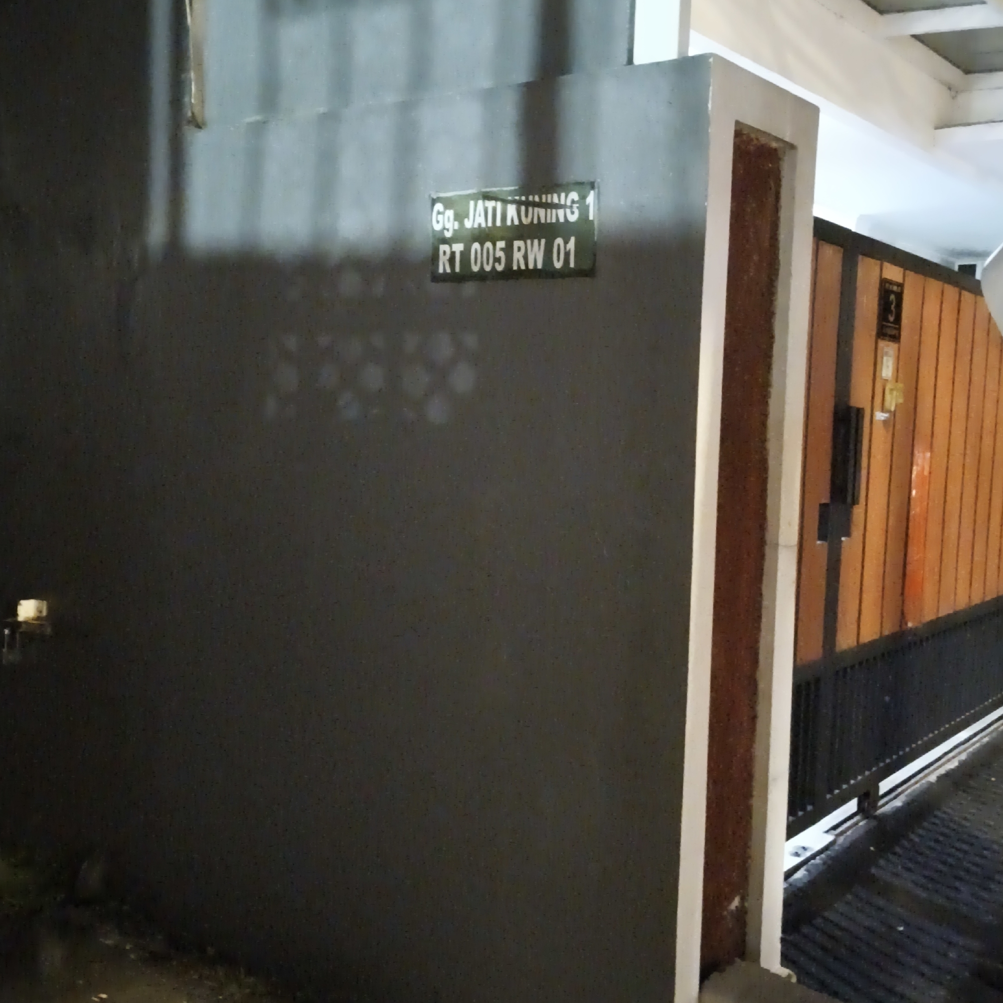 The image shows a street sign in front of a gate in South Jakarta, Indonesia. The sign reads "Gg. JATI KUNING 1 RT 005 RW 01". The gate is made of orange wood and black metal. There is a dark gray wall on the left side of the image. The scene appears to be outdoors at night, with some artificial lighting.
