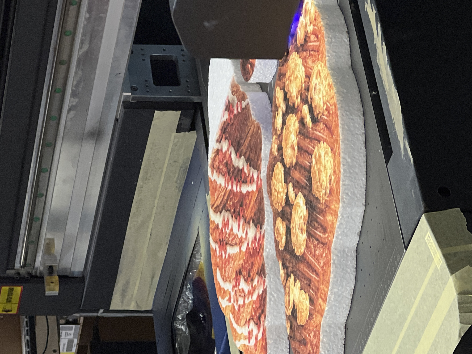 The image displays an industrial setting, likely a print or fabrication shop, focused on a large format printer or cutting machine in operation. In the foreground, two irregular-shaped cutouts, possibly made from foam board or thick paper, are visible. The left cutout features a vibrant pattern of brown and white zigzags, resembling a patterned textile or a stylized animal print. The right cutout, larger and more prominent, depicts a textured brown surface adorned with numerous lighter, round, raised elements, reminiscent of a large croissant or baked good covered in nuts. These cutouts rest on a white, slightly textured surface.
Above the cutouts, a dark, bulky machine head is positioned, suggesting it is either printing or cutting the material. To the left, a metallic linear guide rail with evenly spaced green circular bearings is prominent, indicating precision movement. Further left and in the background, additional metal structural elements of the machine are visible. On the right side of the image, a dark panel features faintly visible measurement markings, including numbers "1 2 3 4 5 6 7 8 9 10," suggesting a ruler or scale. Several pieces of light-colored masking tape are applied to various machine parts. A yellow warning label is partially visible in the bottom left corner. The scene is brightly lit, likely by artificial light, typical of an indoor industrial environment.