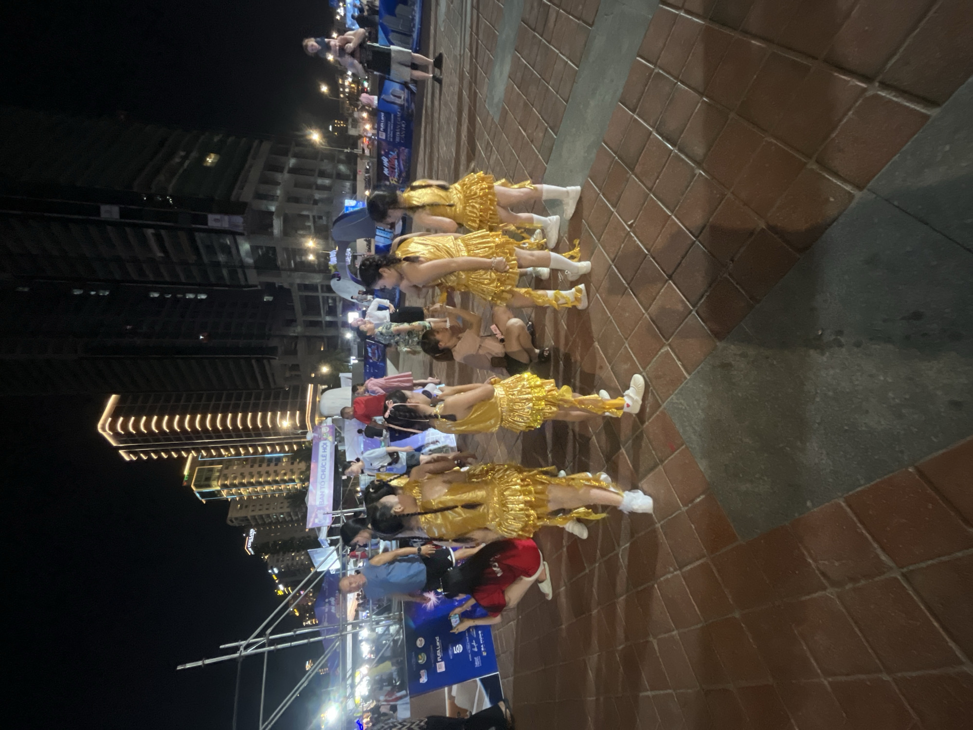 The image depicts an outdoor urban scene at night, likely in Đà Nẵng, Vietnam, given the architectural style and signage that suggests an Asian setting. In the foreground, several young girls dressed in matching gold sequined outfits, likely performers, are gathered. Some are looking at a woman who appears to be adjusting their costumes. The girls are wearing white socks and shoes. The ground is paved with reddish-brown square tiles. In the background, there are tall, modern buildings illuminated with golden lights, characteristic of a city skyline. A large banner with text in what appears to be Vietnamese is visible, possibly advertising an event. Other people are scattered throughout the scene, some walking and others observing. A man is visible on the far right carrying a child on his shoulders. The overall atmosphere appears to be a lively event or gathering.