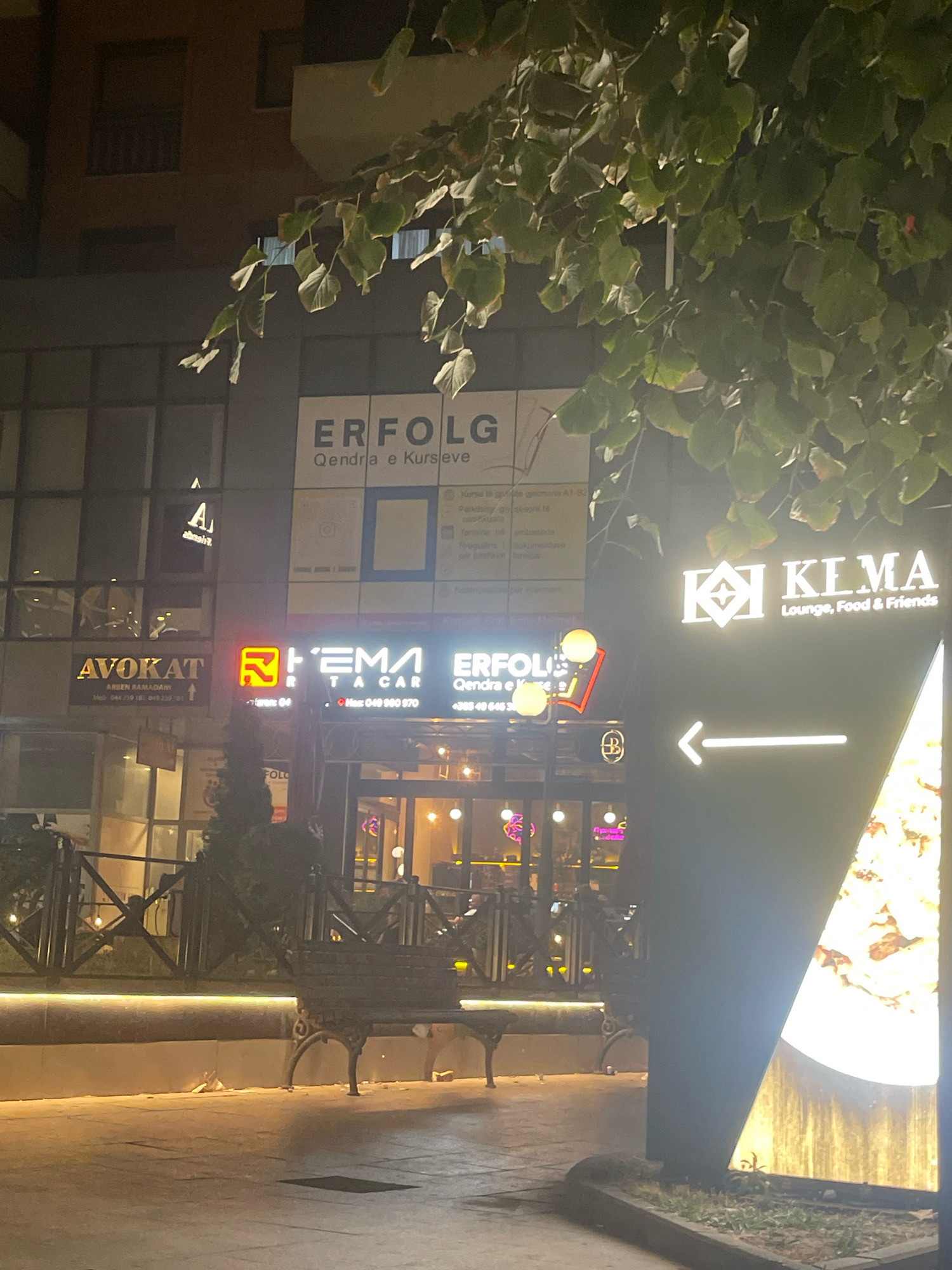 The image depicts a nighttime street scene in Prizren, Kosovo. A tree with green leaves overhangs the frame from the top. In the background, illuminated signs indicate various businesses. One sign reads "ERFOLG Qendra e Kursleve," suggesting an educational center. Another sign displays "AVOKAT," indicating a legal office. Further to the right, a prominent sign for "KIMA Lounge, Food & Friends" is visible, with an arrow pointing left. The scene includes a sidewalk area with a bench, and what appears to be a restaurant or cafe with glowing interior lights. The overall impression is of an urban commercial area at night.