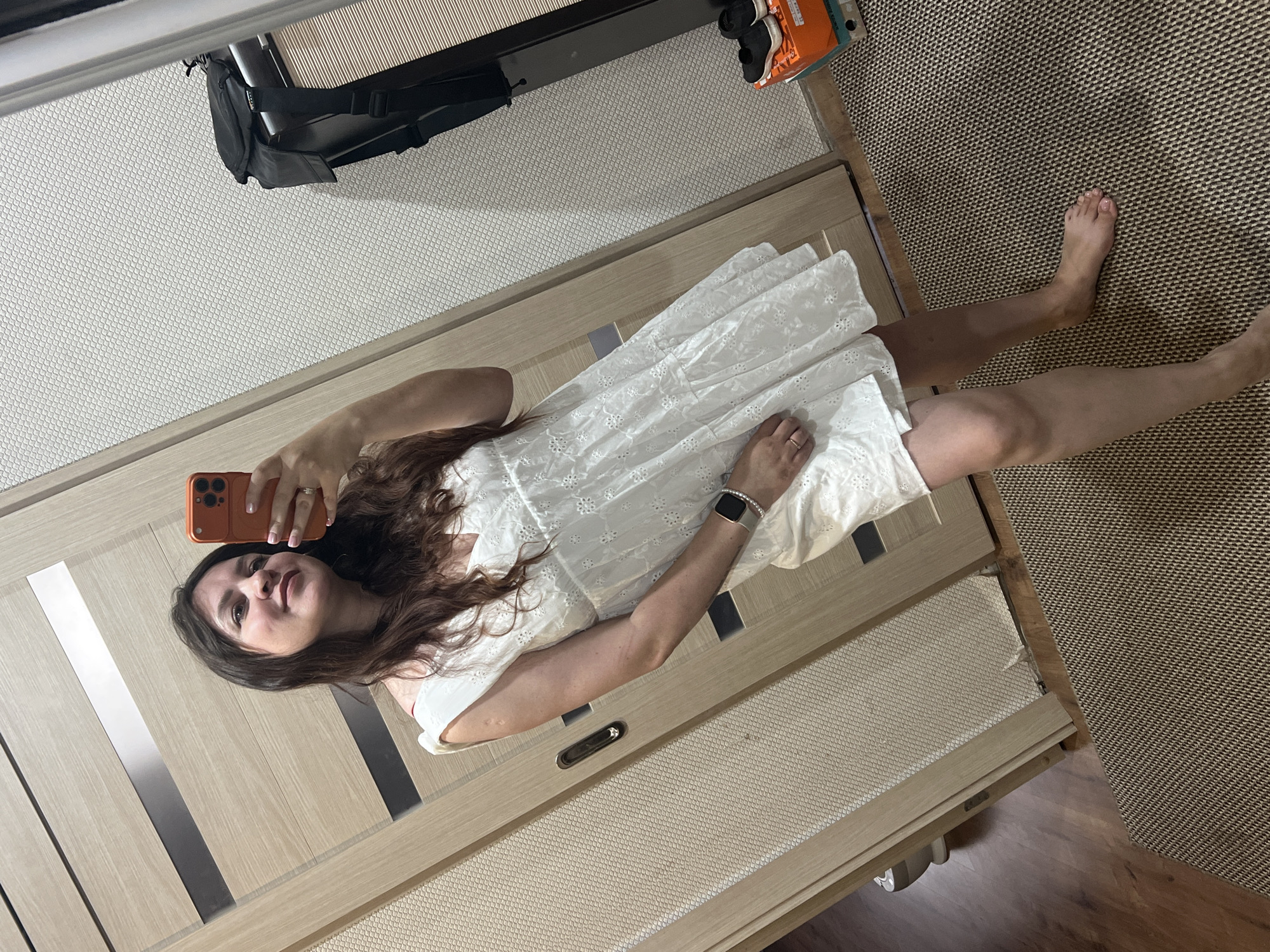 A woman with long, dark hair is taking a selfie in a mirror, holding an orange-cased smartphone in her right hand. She is wearing a white, sleeveless, tiered dress with eyelet embroidery. On her left wrist, she has a silver smartwatch. Her left hand rests on her hip, and a ring is visible on her left ring finger. The background shows a wooden door with horizontal metallic accents and a textured wall. To the right, a black backpack is visible on a shelf, with a cardboard box containing black sneakers below it. The floor is covered with a woven rug, and a wooden floor is visible to the left. The setting appears to be an indoor domestic environment, possibly a hallway or entryway. There is no discernible indication of the location being in Noviy, Russia.