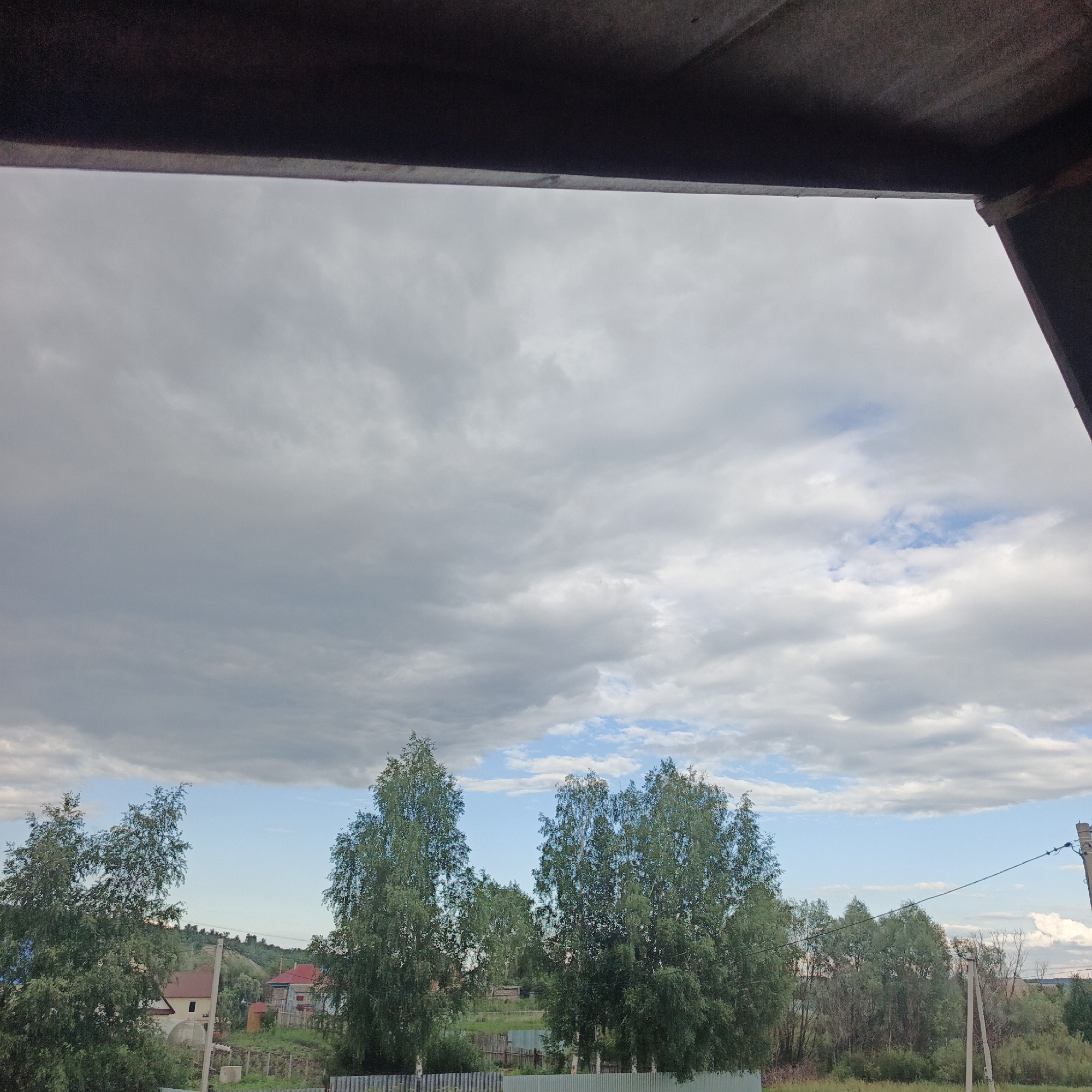 This image depicts a cloudy sky over a rural landscape in Podlubovo, Russia. The foreground is partially obscured by a dark, overhanging structure, possibly a roof or canopy, suggesting the photo was taken from indoors or a covered outdoor area.
The sky is filled with a mix of gray and white clouds, with patches of blue sky visible, indicating a partly cloudy or overcast day. The clouds are varied in texture, with some appearing dense and dark, while others are lighter and more scattered.
Below the sky, a line of trees, primarily deciduous with green foliage, stretches across the middle ground. Among the trees, some houses with red roofs are visible, along with fences and what appear to be utility poles and wires, suggesting a residential area within a rural setting. The vegetation is green and lush, typical of a warmer season. In the background, rolling hills are visible, covered with trees.
There is no discernible activity or event taking place; the scene appears to be a still landscape. The overall mood is somewhat somber due to the overcast sky, but the presence of green foliage and patches of blue sky offers a sense of natural beauty and calm. The time of day is likely daytime, given the lighting conditions, but the cloud cover makes it difficult to determine precisely. No text is visible in the image.