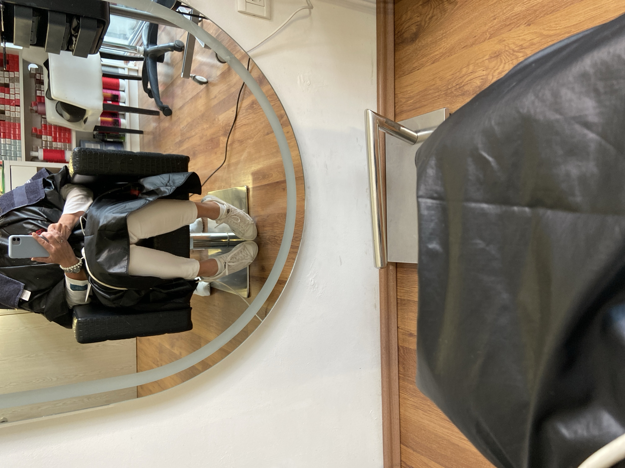 Captured in an indoor setting, this image presents a reflection in an illuminated, circular mirror. A person wearing white trousers and sneakers is seated in a salon chair, a dark cape draped over their shoulders, visible in the mirror. The person is holding a mobile phone. The background of the reflection reveals a portion of the salon with a shelf of hair dye products and a stylist's station. In the foreground, outside the reflection, is a closed door with a silver handle. A dark, draped salon cape partially covers the door and obscures part of the view. The location, Budva, Montenegro, is only inferred as a possible location.