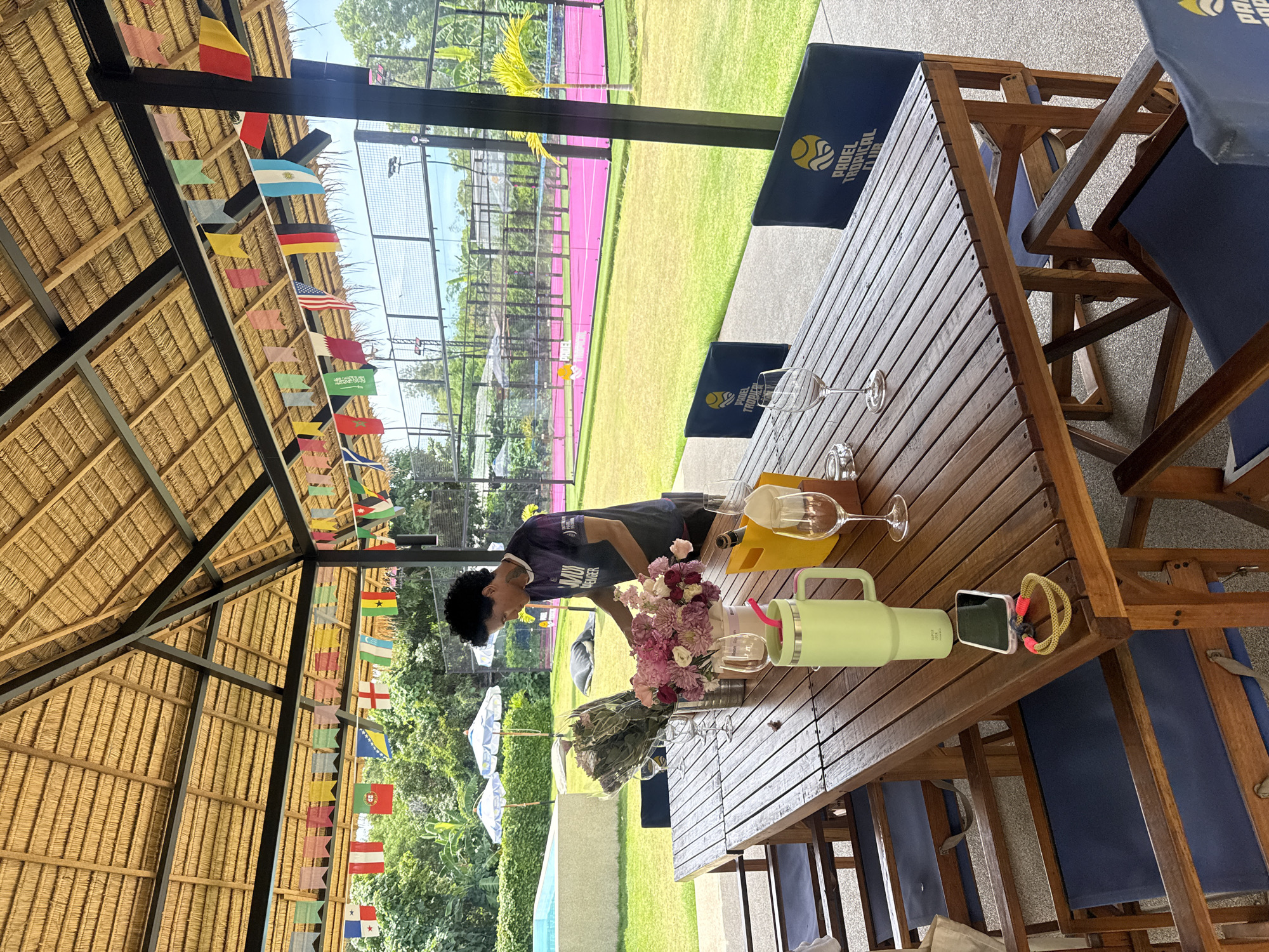 A man is arranging flowers on a wooden table at an outdoor covered seating area with a thatched roof. The table is laden with floral arrangements, wine glasses, a yellow cooler, and a large green insulated tumbler with a pink straw. A smartphone and a coiled yellow rope are on the table. In the background, a pink paddle tennis court is visible, along with lush greenery, umbrellas, and a swimming pool. The area is decorated with a string of international flags. The man is wearing a dark t-shirt with a logo. The overall setting appears to be a resort or club in บ้านบางรักษ์, Thailand, likely at a Padel Tropical Club, given the signage on the chairs.