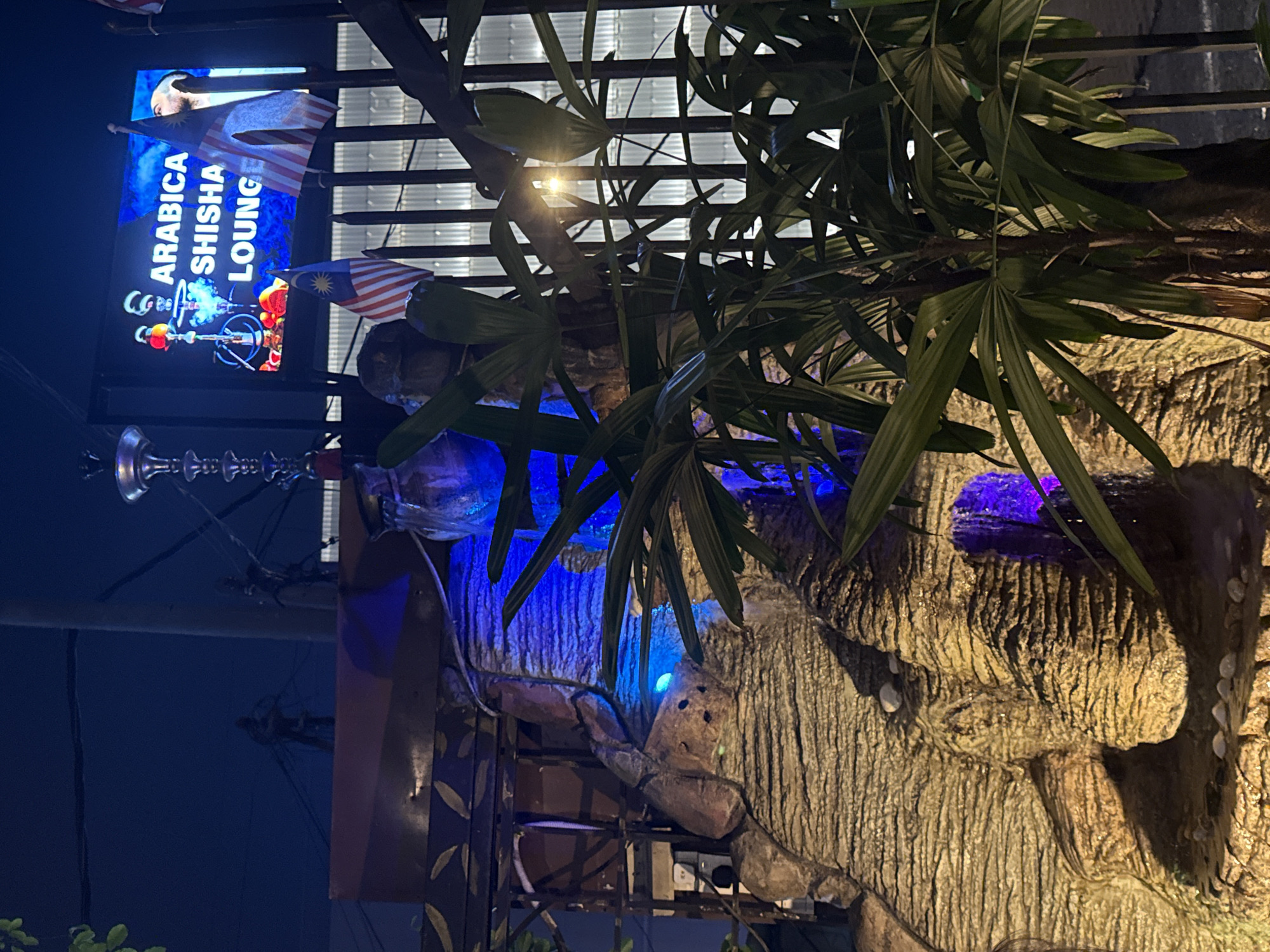 This night-time image captures the exterior of an establishment called "ARABICA SHISHA LOUNGE" in Kuala Lumpur, Malaysia. A large, vertically oriented illuminated sign, primarily lit with blue light, prominently displays "ARABICA SHISHA LOUNGE" in white capital letters. The sign features a graphic of a shisha pipe emitting smoke, with a subtle image of a person at the top.
Two Malaysian flags are visible; one is attached near the top of the main sign, and another smaller one is further to the right, confirming the location. The scene is characterized by dense, dark green foliage, possibly palm leaves, which partially obscure the background on the right and top.
Below and to the right of the sign, a textured, rock-like structure or wall is illuminated with striking blue lights, creating a distinctive ambiance. Smaller white or light-colored accents are also visible on this structure. A tall, decorative shisha pipe with intricate silver detailing is partially visible on the far left, suggesting the lounge's theme. The overall lighting scheme, with the dark sky and vibrant blue artificial lights, indicates it is evening or night. The setting appears to be an inviting outdoor or semi-outdoor space for the shisha lounge.