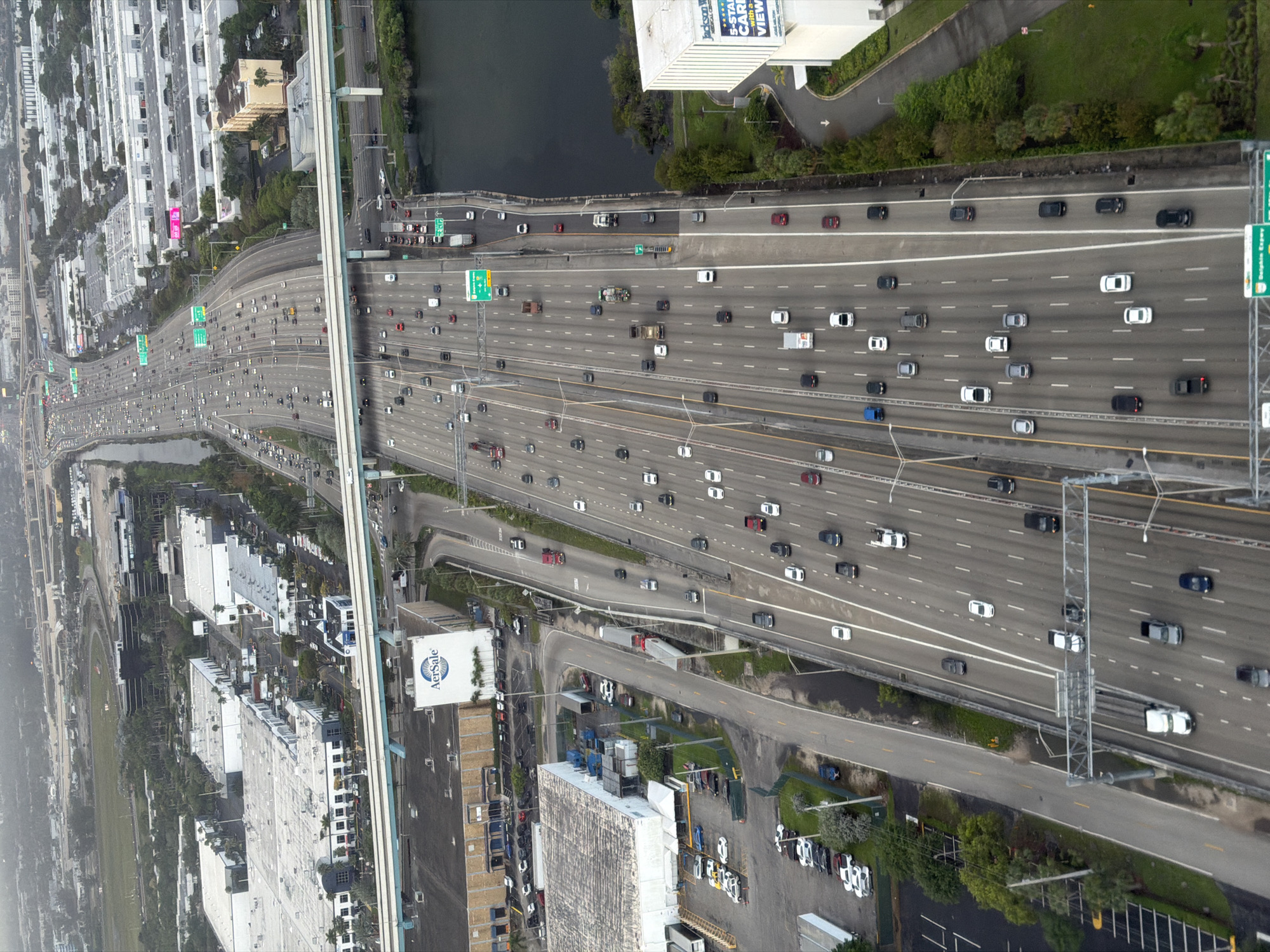 The image depicts a high-angle view of a multi-lane highway with heavy traffic, likely an interstate or major expressway. Numerous vehicles, mostly cars and trucks, are visible on the road. The highway is surrounded by urban development, including buildings, parking lots, and some green spaces. A body of water borders the highway on one side, while an elevated guideway or monorail structure runs parallel to it. Various road signs are visible above the highway lanes. Based on the architecture, landscape, and roadway design, the image likely captures a scene from Miami, Florida, United States.