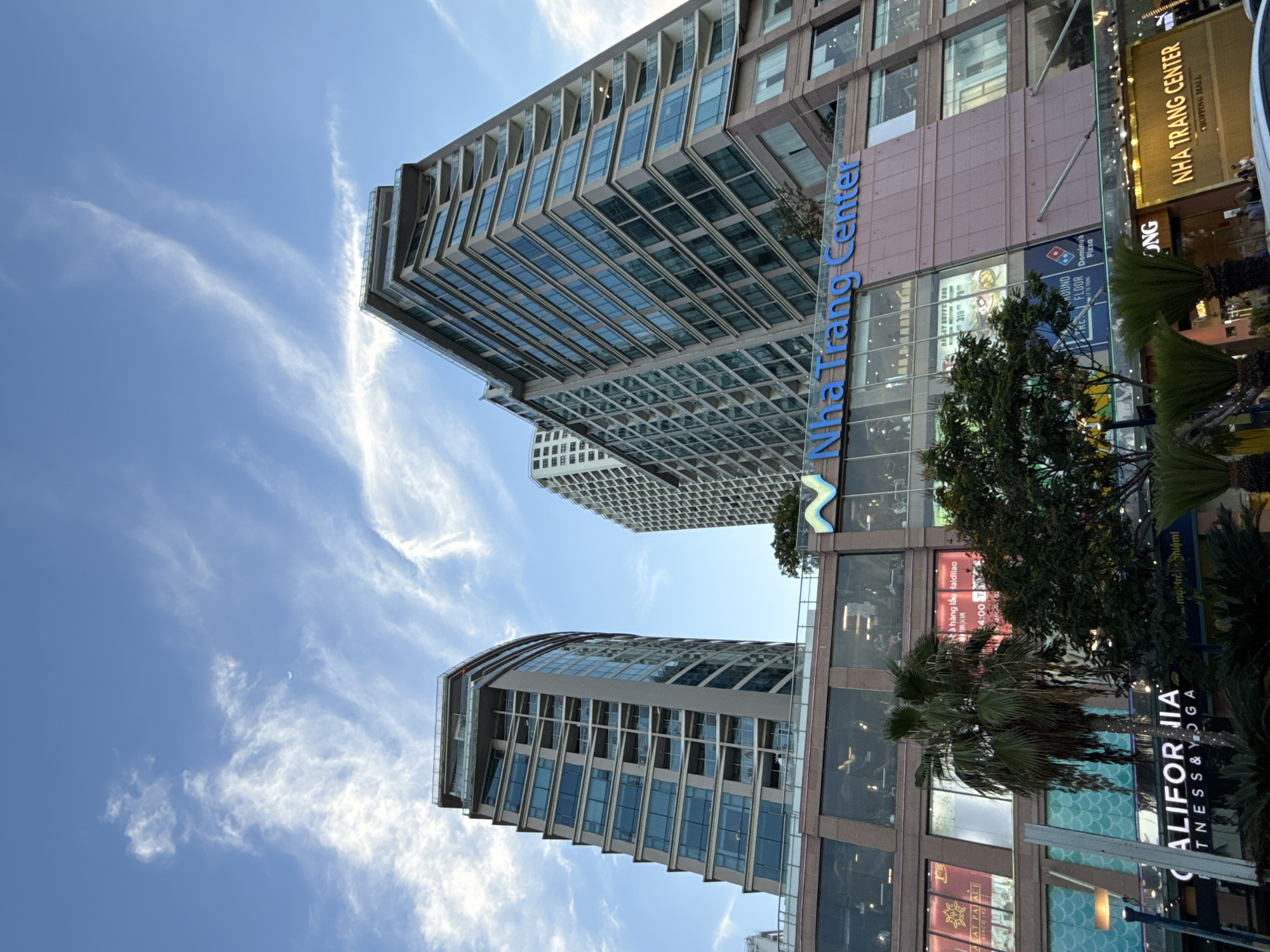 A low-angle exterior shot captures the Nha Trang Center, a modern shopping mall and apartment complex, against a bright blue sky dotted with wispy white clouds. The building features multiple stories of glass-fronted apartments with balconies, interspersed with floors of retail spaces. The ground level is occupied by shopfronts displaying various advertisements, including one for "California FITNESS & YOGA." Palm trees and other lush greenery are visible in front of the lower floors, adding a tropical ambiance. The "Nha Trang Center" logo is prominently displayed in blue lettering on the facade. The scene appears to be located in the city of Nha Trang, Vietnam.